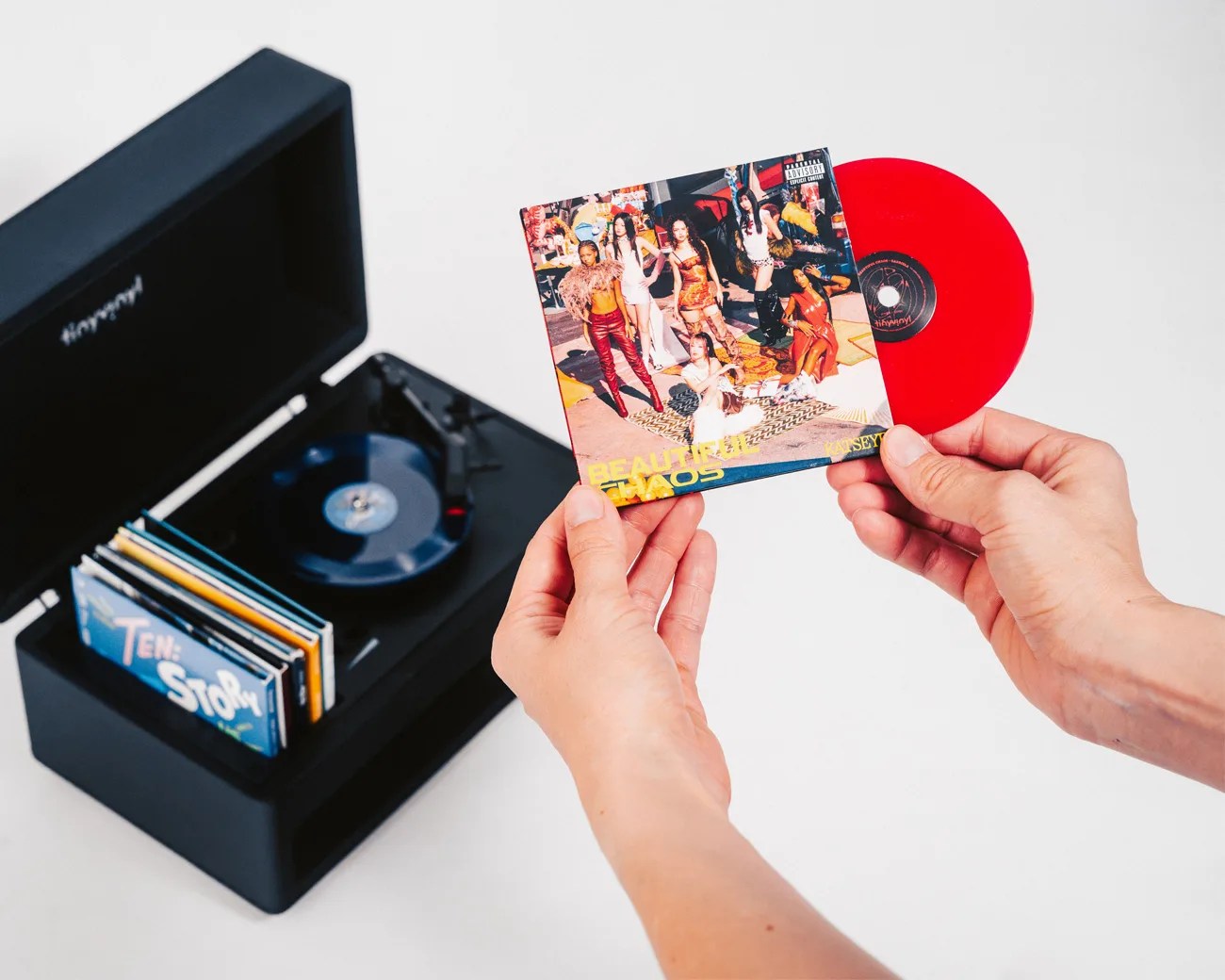 Hands holding a red vinyl record and its colorful album cover with a group photo, next to a black record player with other albums.