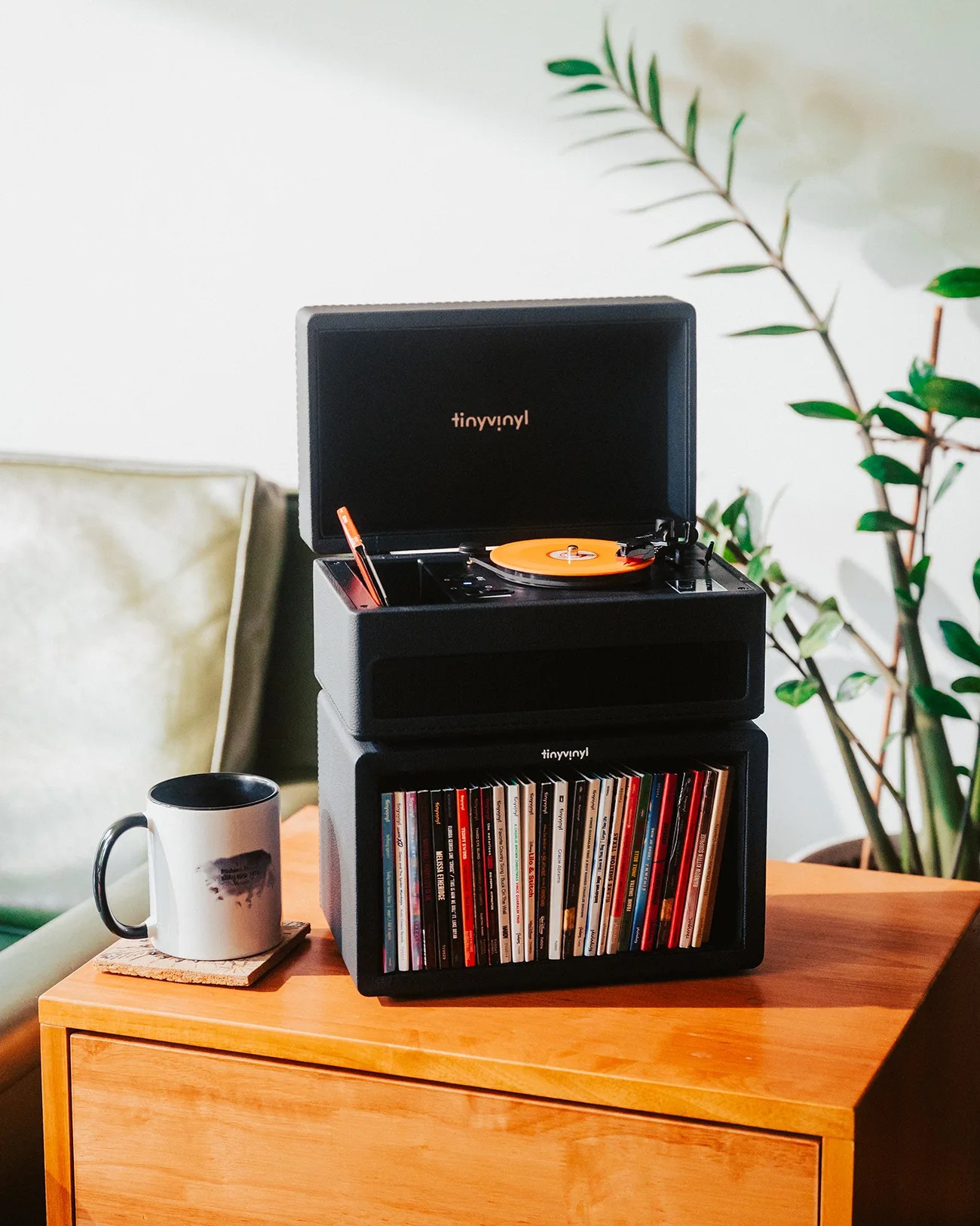 Black TinyVinyl record player with orange vinyl on top of a matching black record storage unit filled with records on a wooden table.