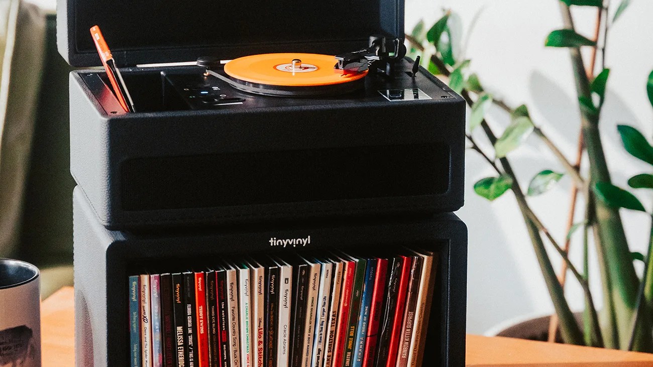Black Tinyvinyl turntable with orange vinyl record on top of a matching black storage unit filled with vinyl records.