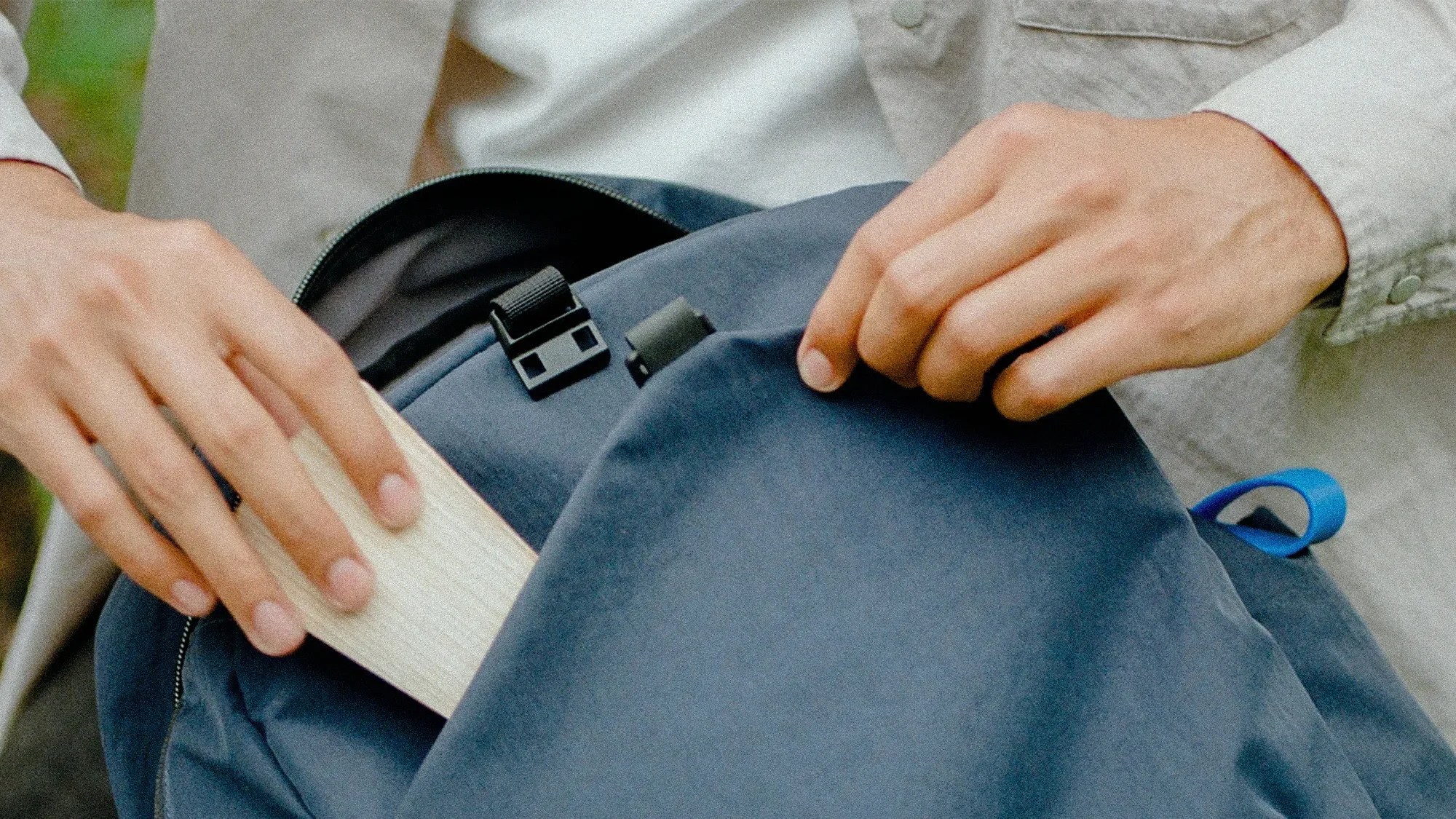 Hands placing a light wooden object into a partially open dark blue backpack with black buckle and blue loop.