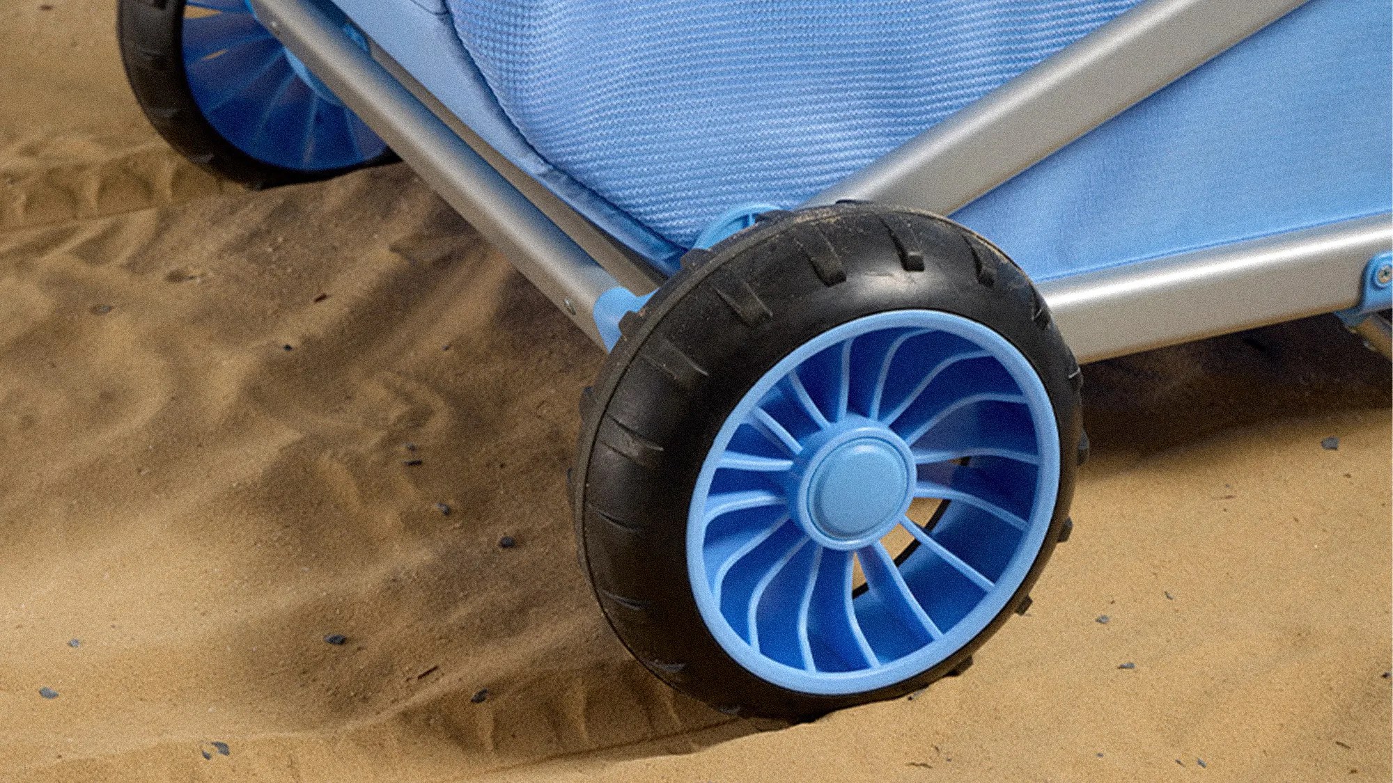 Close-up of black and blue plastic wheels attached to a light blue fabric and metal frame on sand.