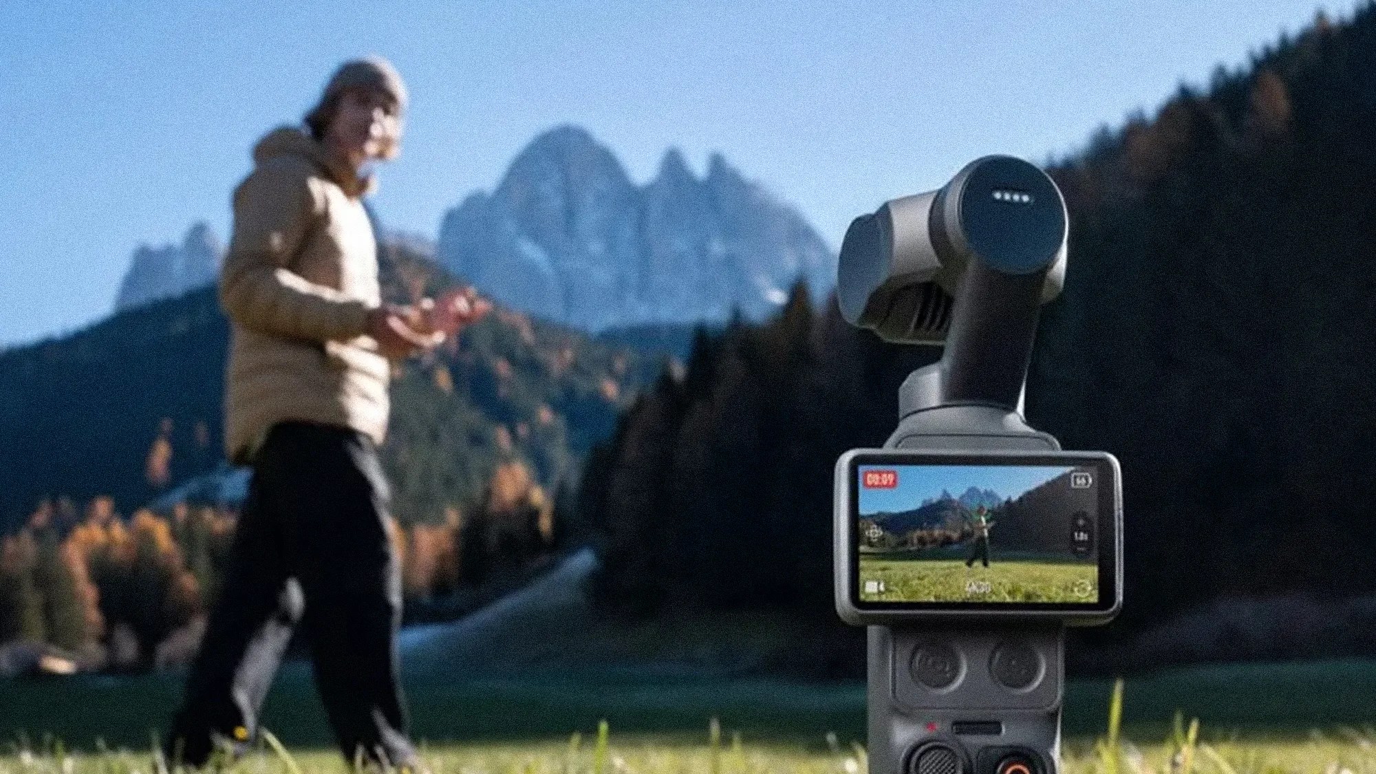 Handheld gray camera gimbal with screen showing a person in a field, mountains and trees in the background.