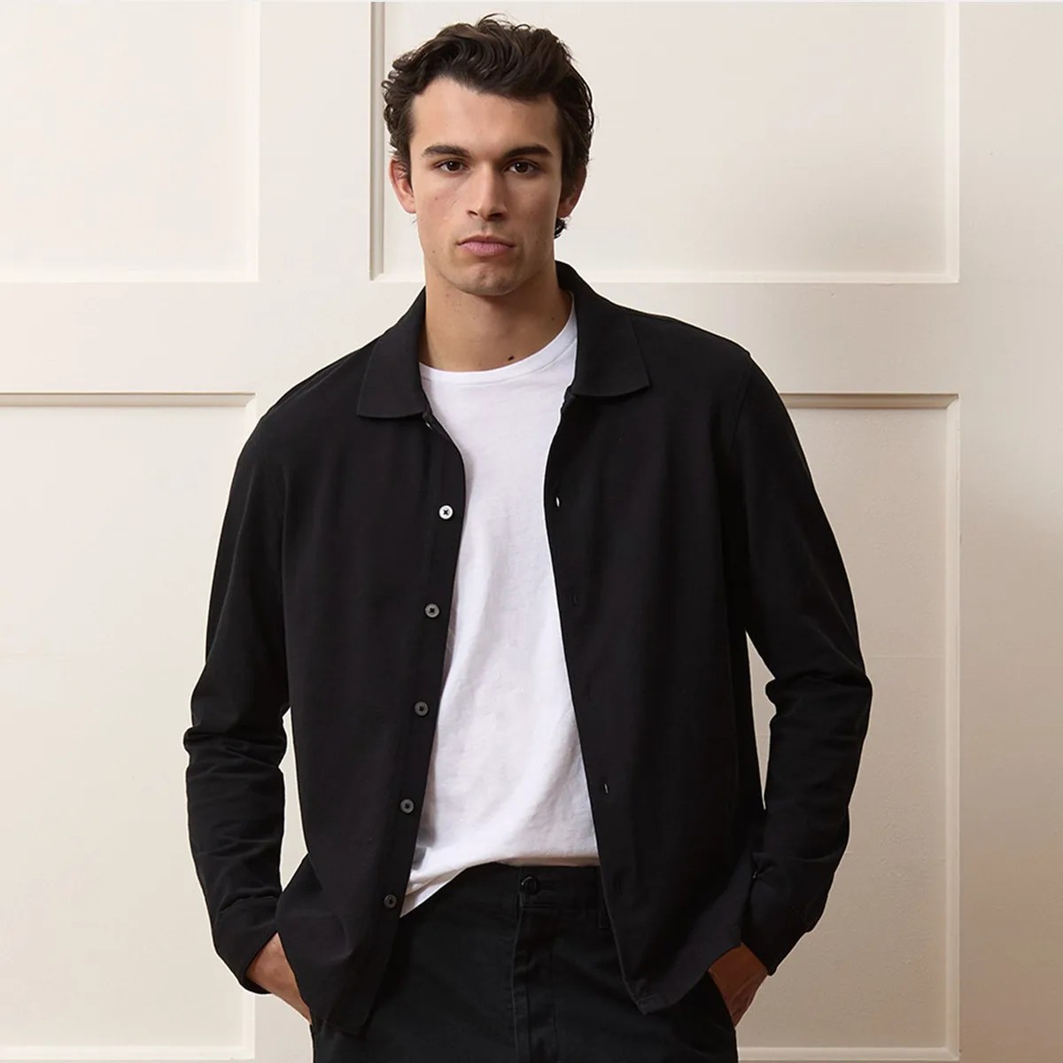 Man wearing a black button-up shirt over a white t-shirt with hands in pockets.