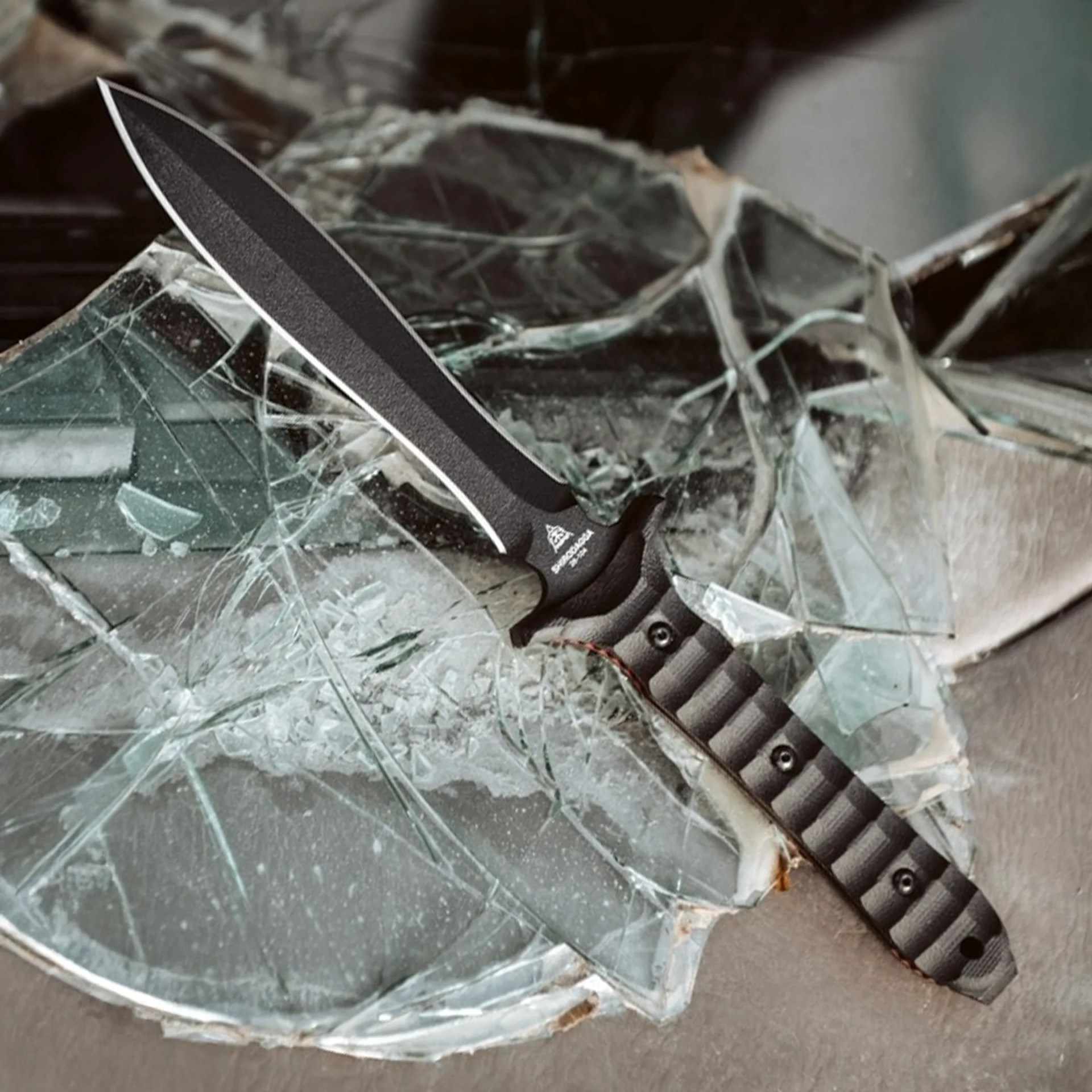 Black tactical knife with textured grip handle stabbed into shattered glass on a concrete surface.