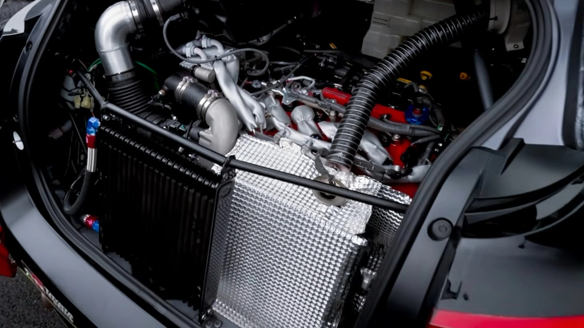 Close-up of a car engine bay showing a silver heat shield, black hoses, and various engine components.