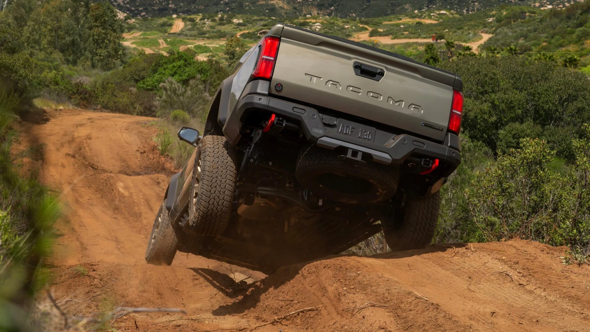 Gray Toyota Tacoma truck with off-road tires driving on a dirt trail in a green hilly area.