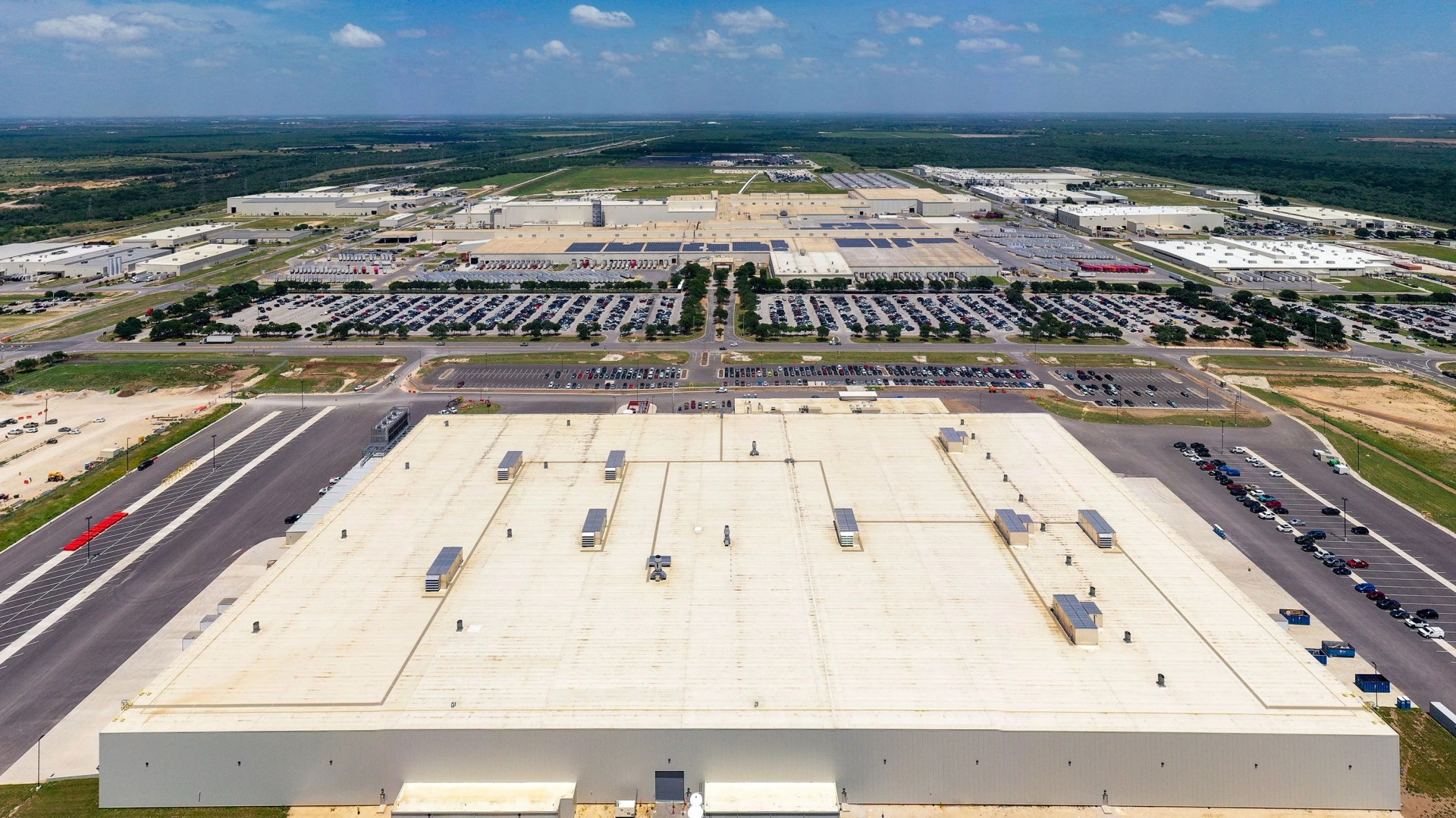 Aerial view of a large industrial warehouse complex with multiple parking lots filled with cars and surrounding greenery.