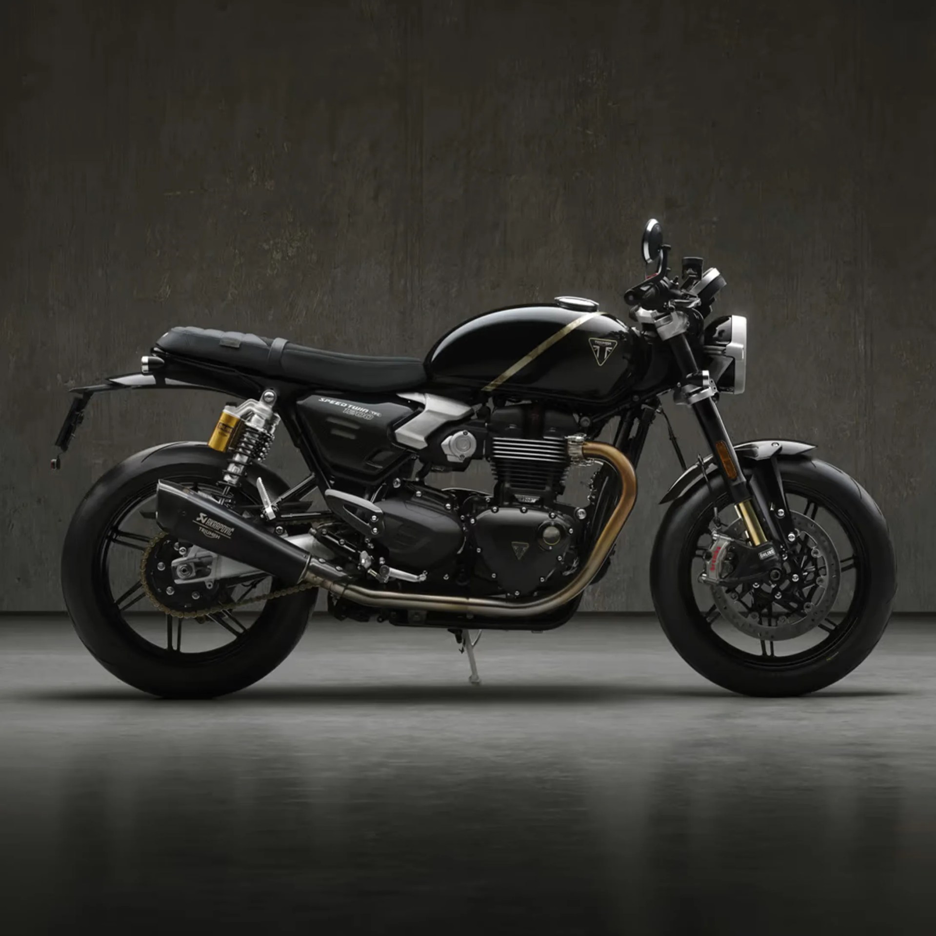 Black Triumph Speed Twin motorcycle with gold exhaust pipe and yellow rear suspension against a dark textured wall.