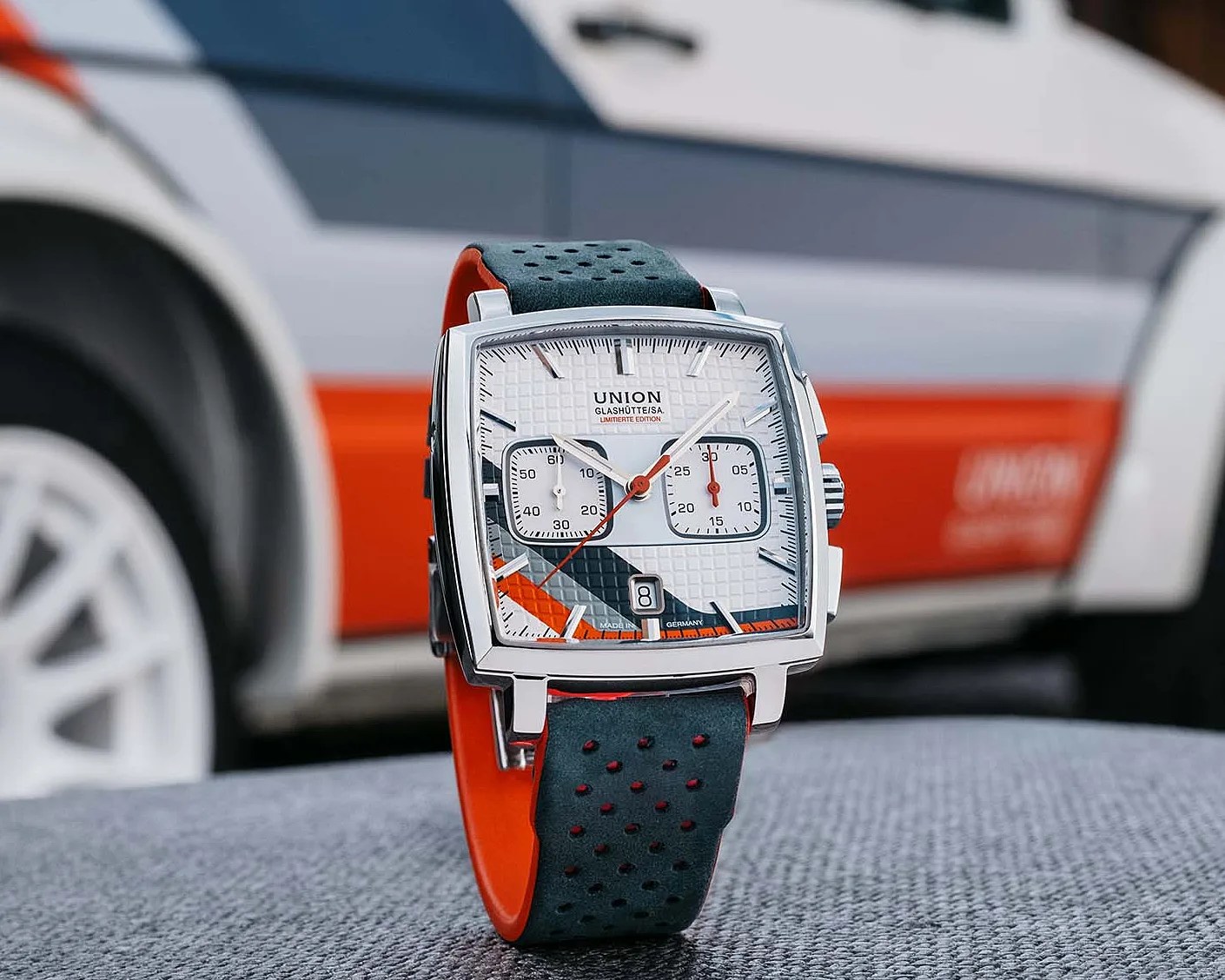 Square silver wristwatch with white textured dial, black and orange accents, and perforated black strap with orange underside.