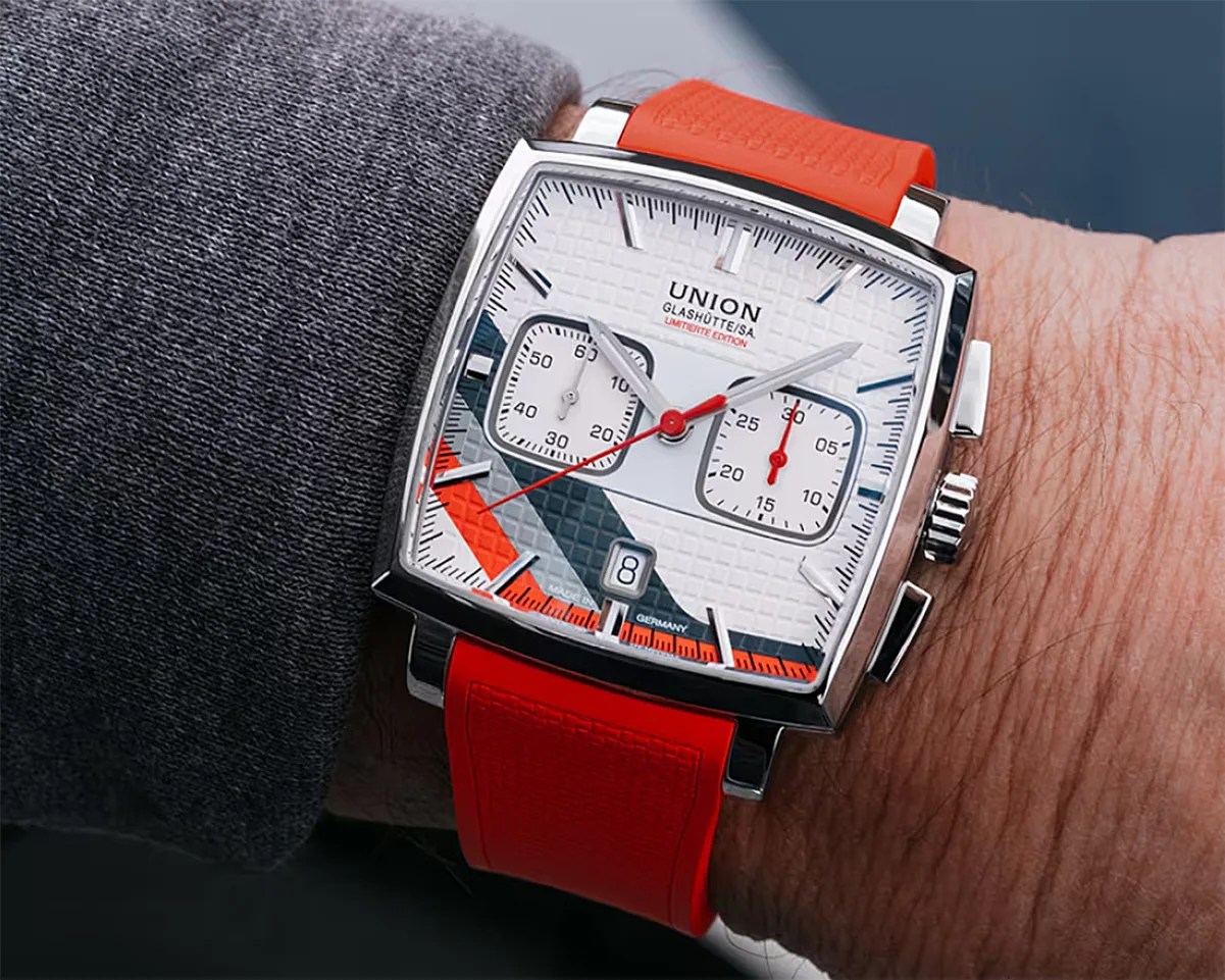 Square silver wristwatch with white textured dial, red and black accents, and bright red strap worn on a wrist.