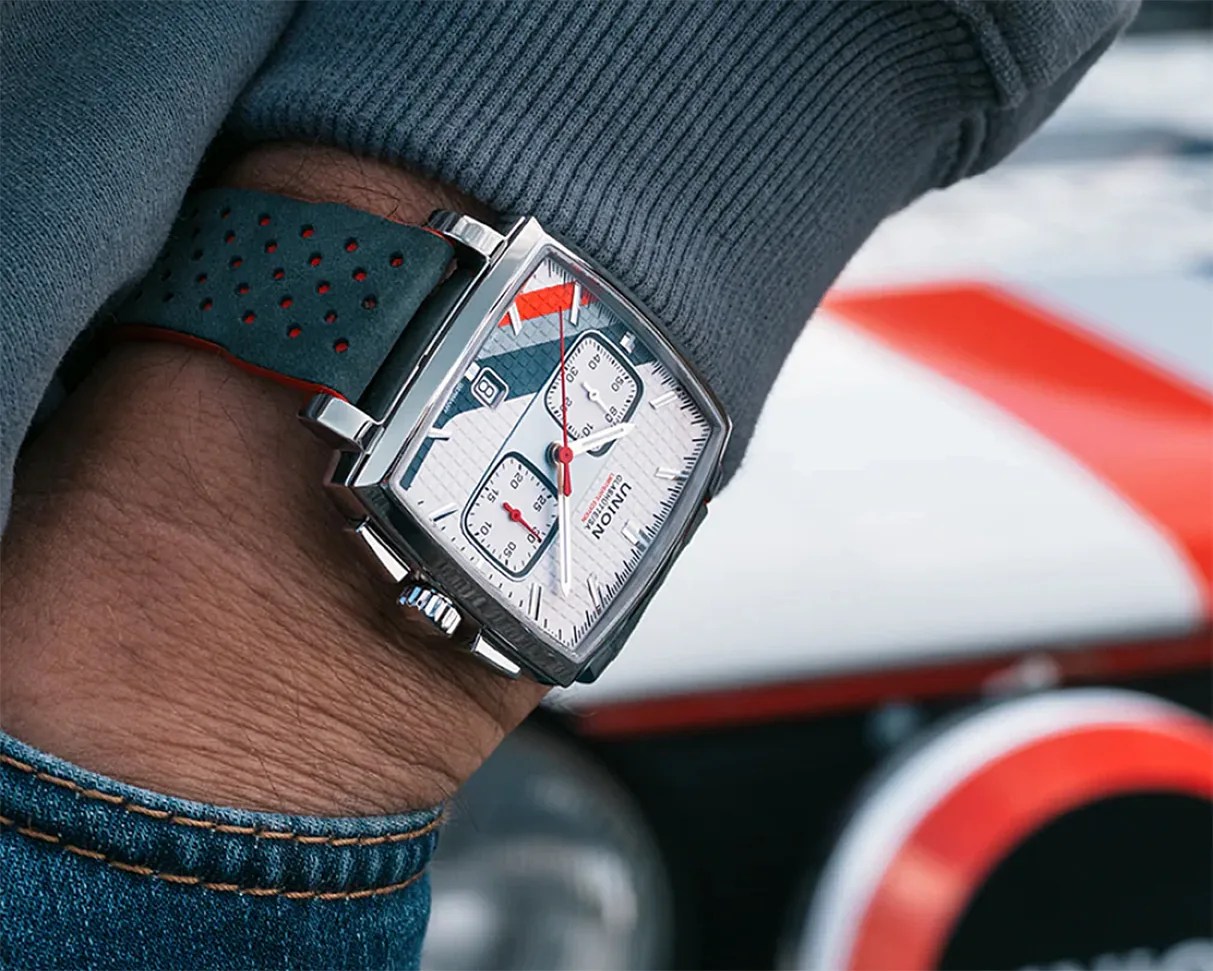 Square silver wristwatch with white dial, red and black accents, and perforated black strap worn on a wrist.