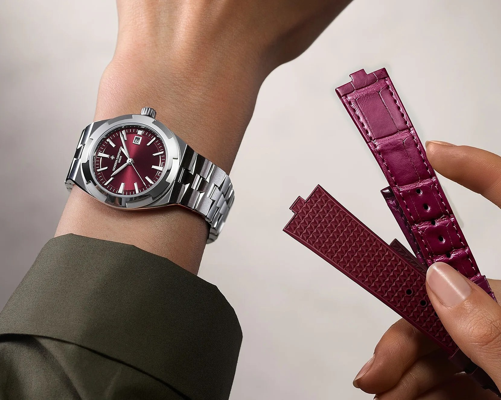 Silver metal wristwatch with a burgundy dial worn on a wrist, next to two interchangeable burgundy watch straps held in a hand.
