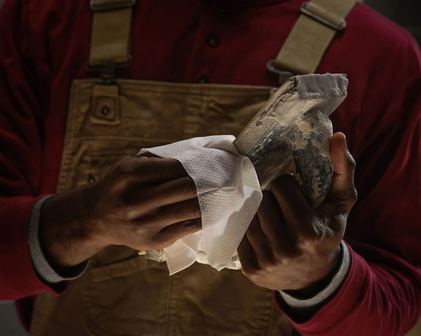 Hands wiping a worn ballet pointe shoe with a white cloth, person wearing brown overalls and a red shirt.