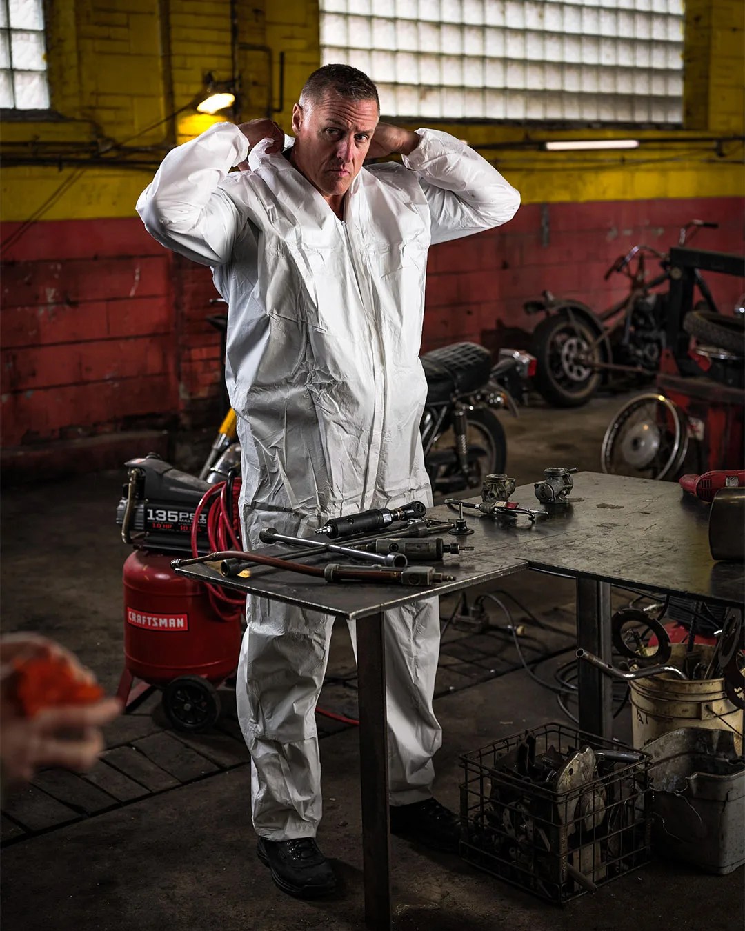 Man putting on a white protective coverall in a workshop with tools on a metal table.