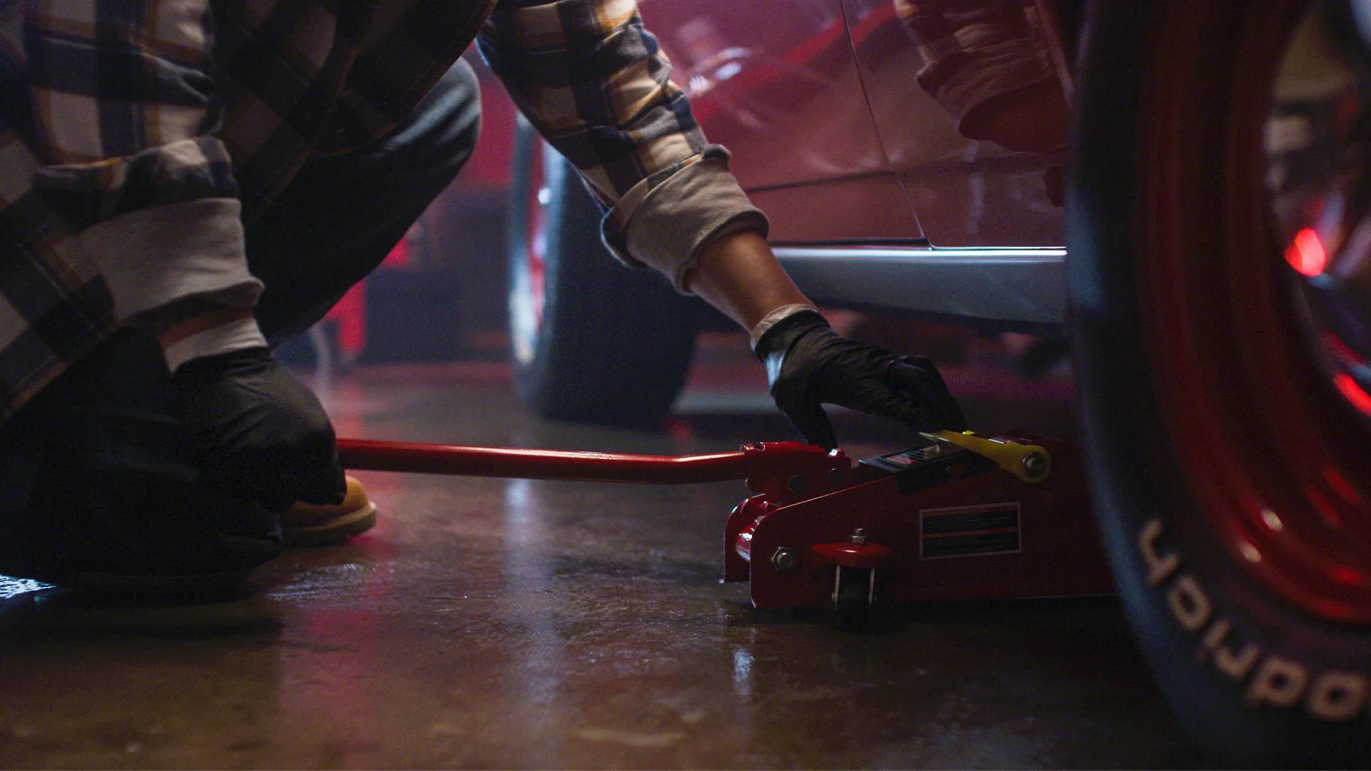 Person wearing black gloves and a plaid shirt operating a red hydraulic floor jack under a red car.