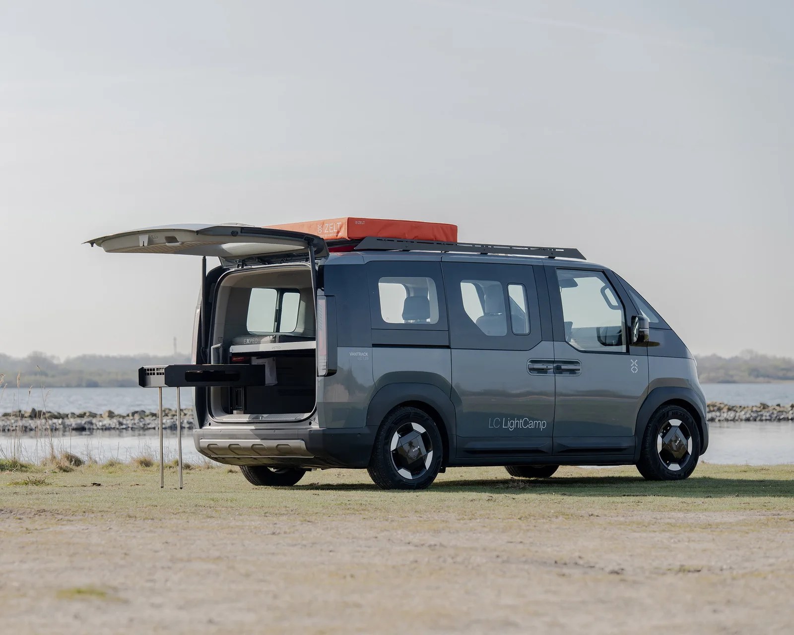 Gray Kia Vantrack LC LightCamp camper van with rear hatch open and pull-out kitchen setup by a lakeside.