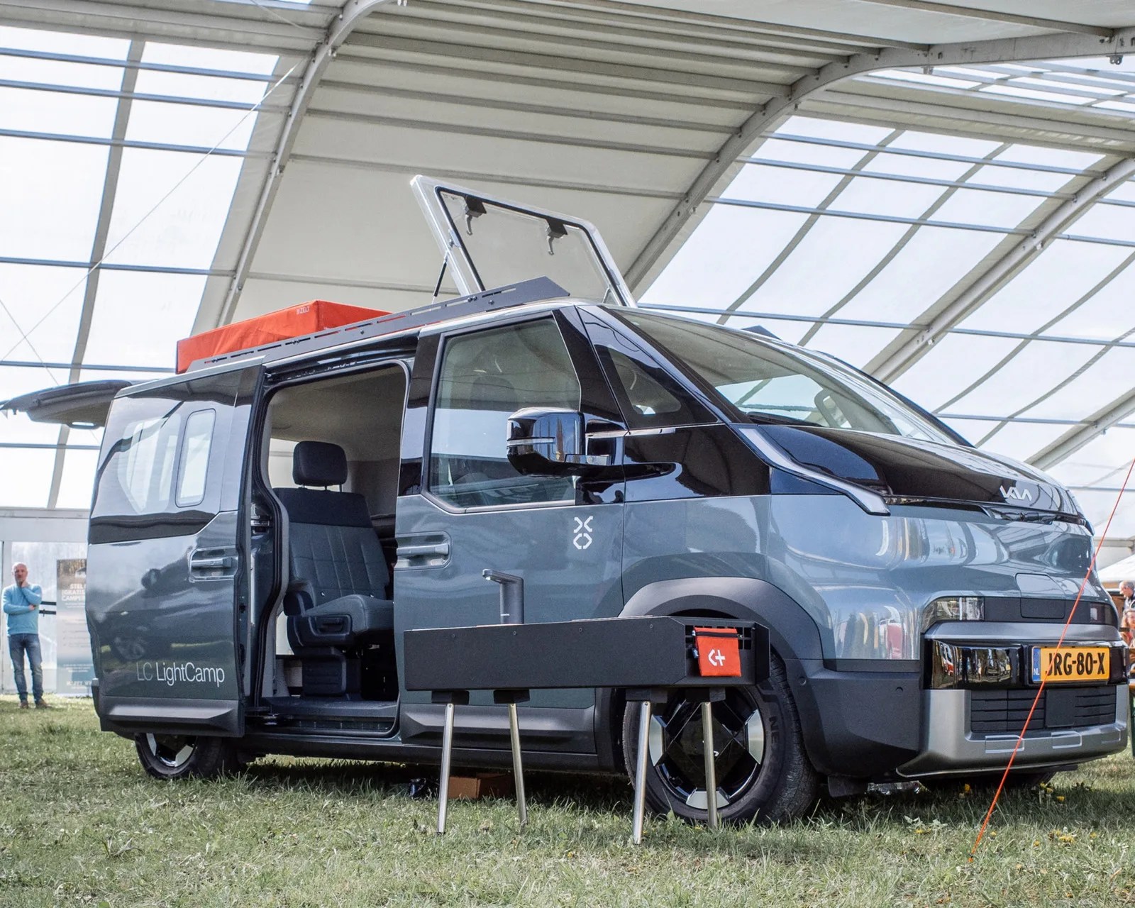 Gray and black Kia LightCamp camper van with sliding door open and roof hatch raised, parked on grass indoors.