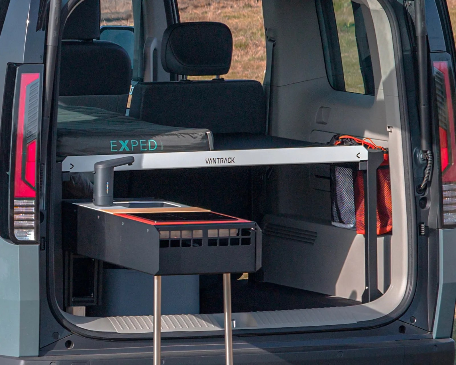 Rear view of a van with a black Exped mattress on a Vantrack bed frame and a portable stove inside.