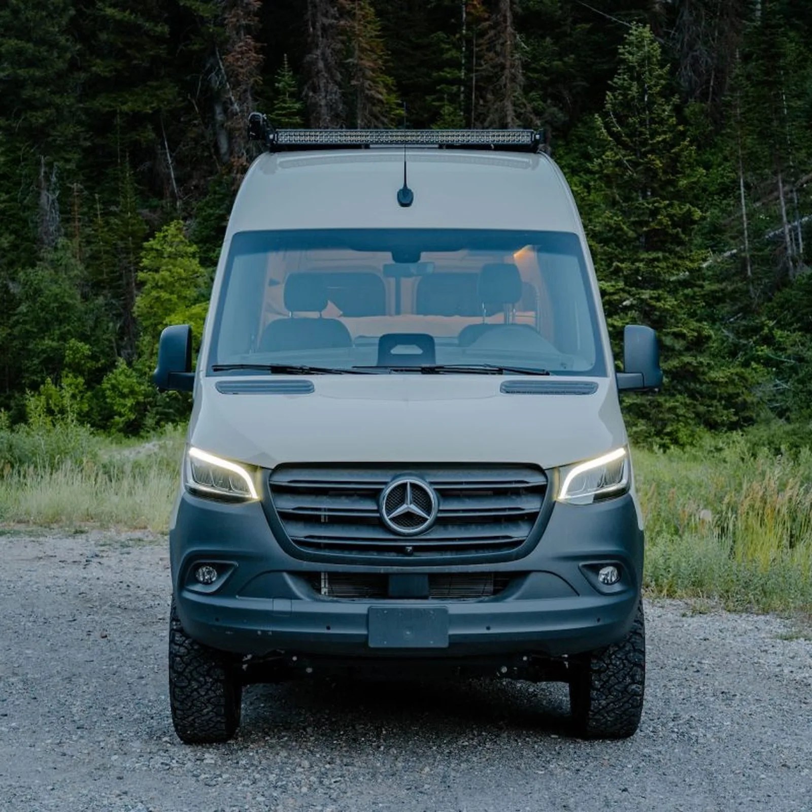 White Mercedes-Benz Sprinter van with LED light bar on roof parked on gravel road with forest background.