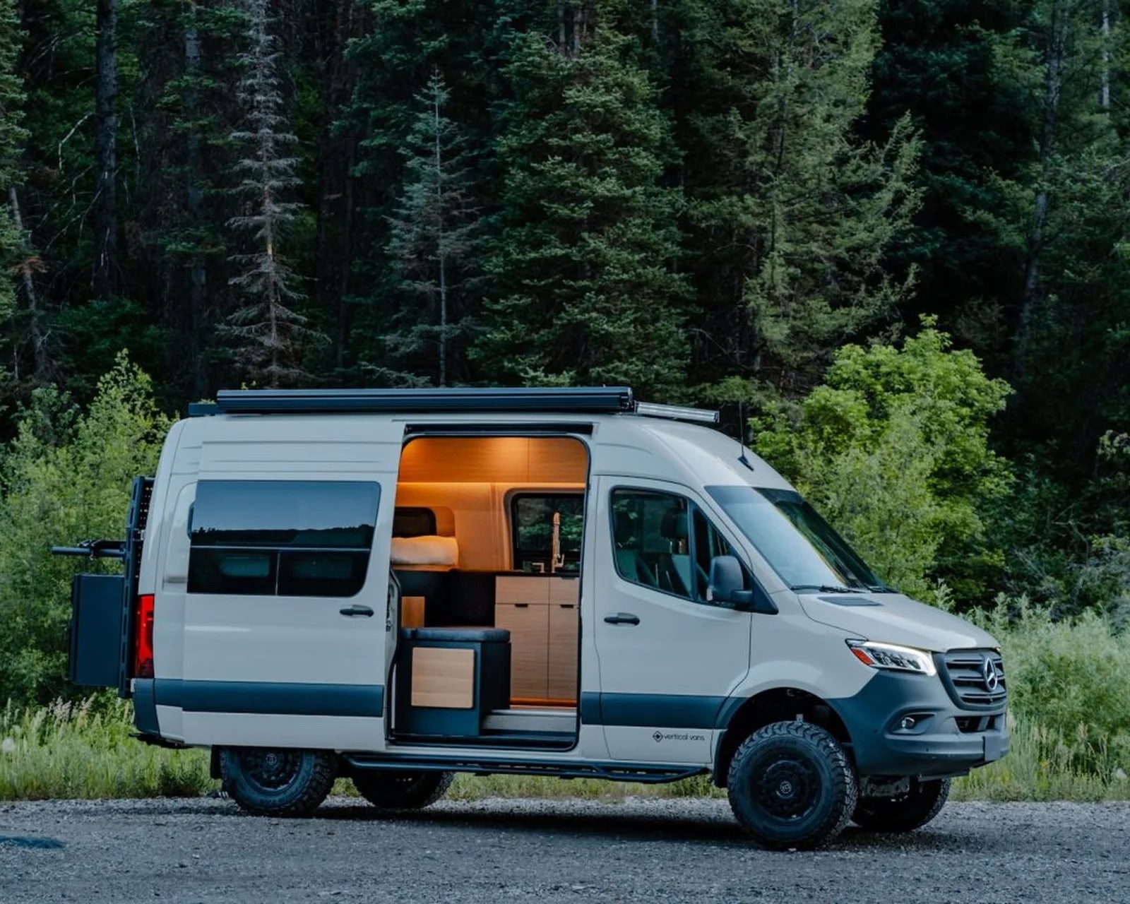 White Mercedes camper van with sliding door open showing wooden interior, parked on gravel near forest.