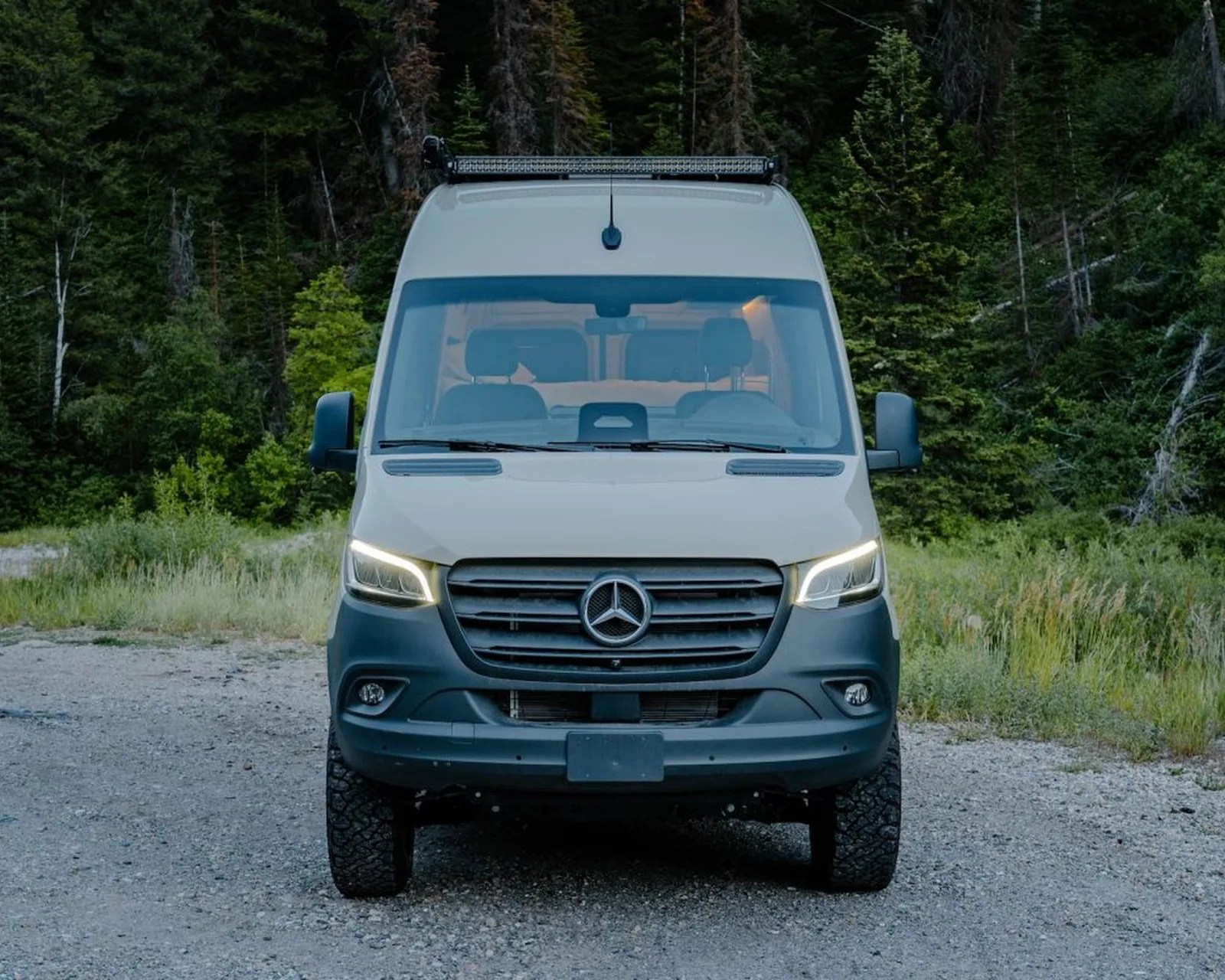Front view of a white Mercedes-Benz van with LED headlights on a gravel road surrounded by dense green forest.