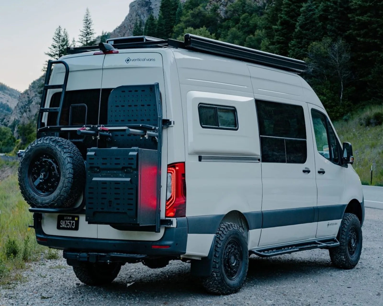 White camper van with off-road tires, roof rack, rear ladder, spare tire, and side window awning parked on a gravel road.
