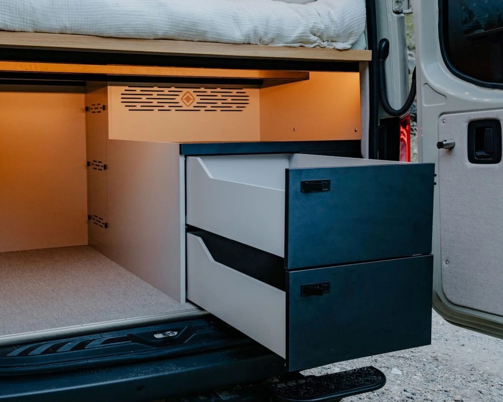 Two-tone sliding storage drawers extended from the side of a camper van under a bed platform.