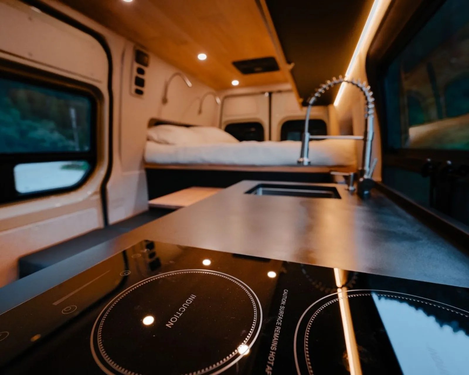 Black induction cooktop with two burners in a compact camper van kitchen with a bed in the background.