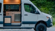 White camper van with open sliding door showing wood-finished kitchen cabinets and black faucet inside.