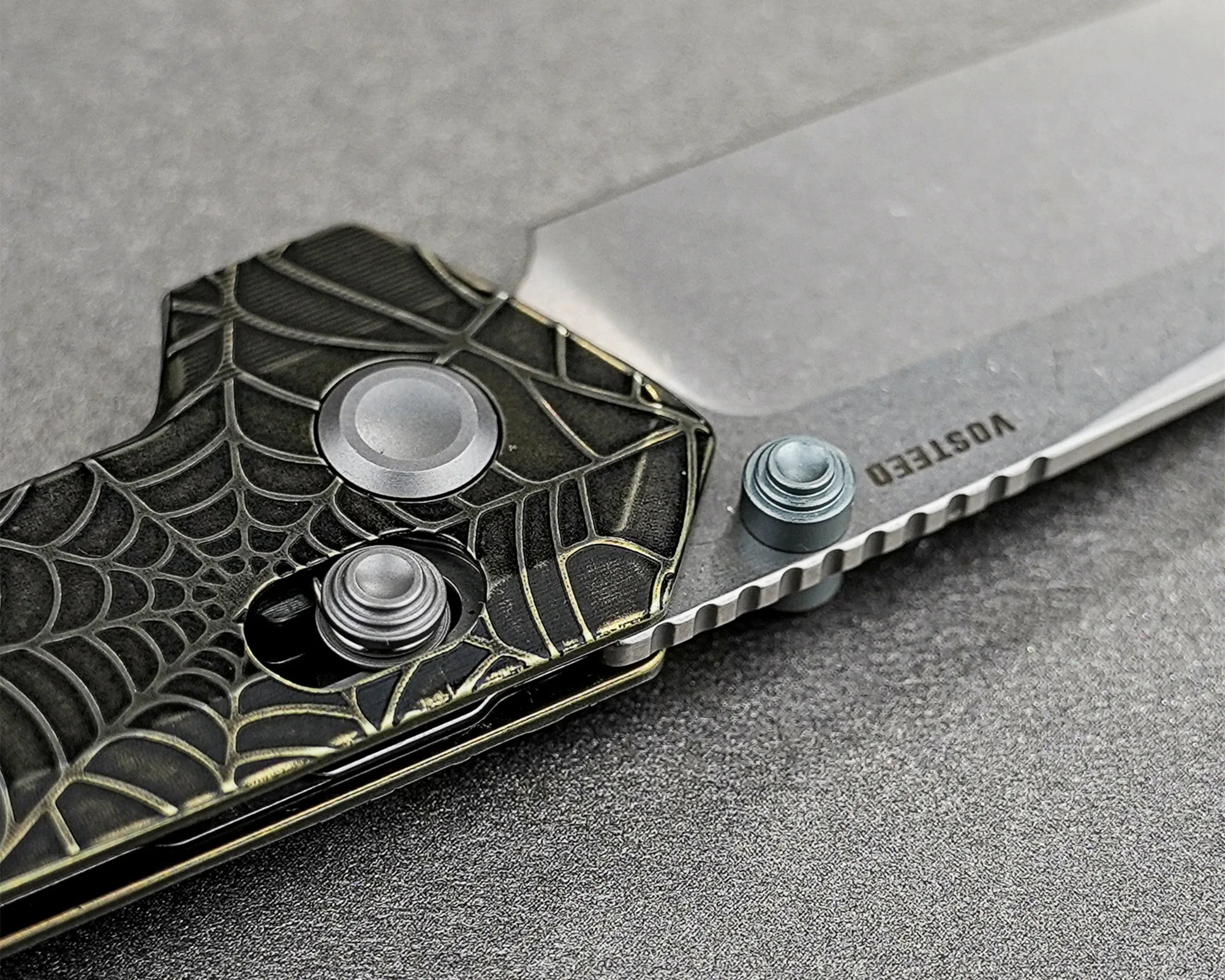 Close-up of a folding knife with a spider web patterned handle and a blade engraved with "VOSTEED.