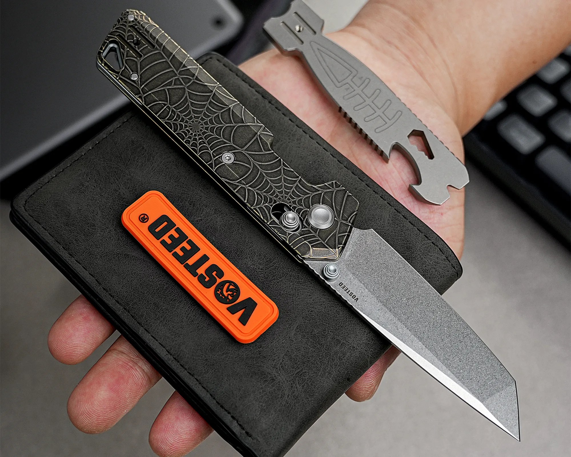 Black leather wallet with an orange Vosteed patch, a spider web patterned folding knife, and a gray multi-tool held in a hand.