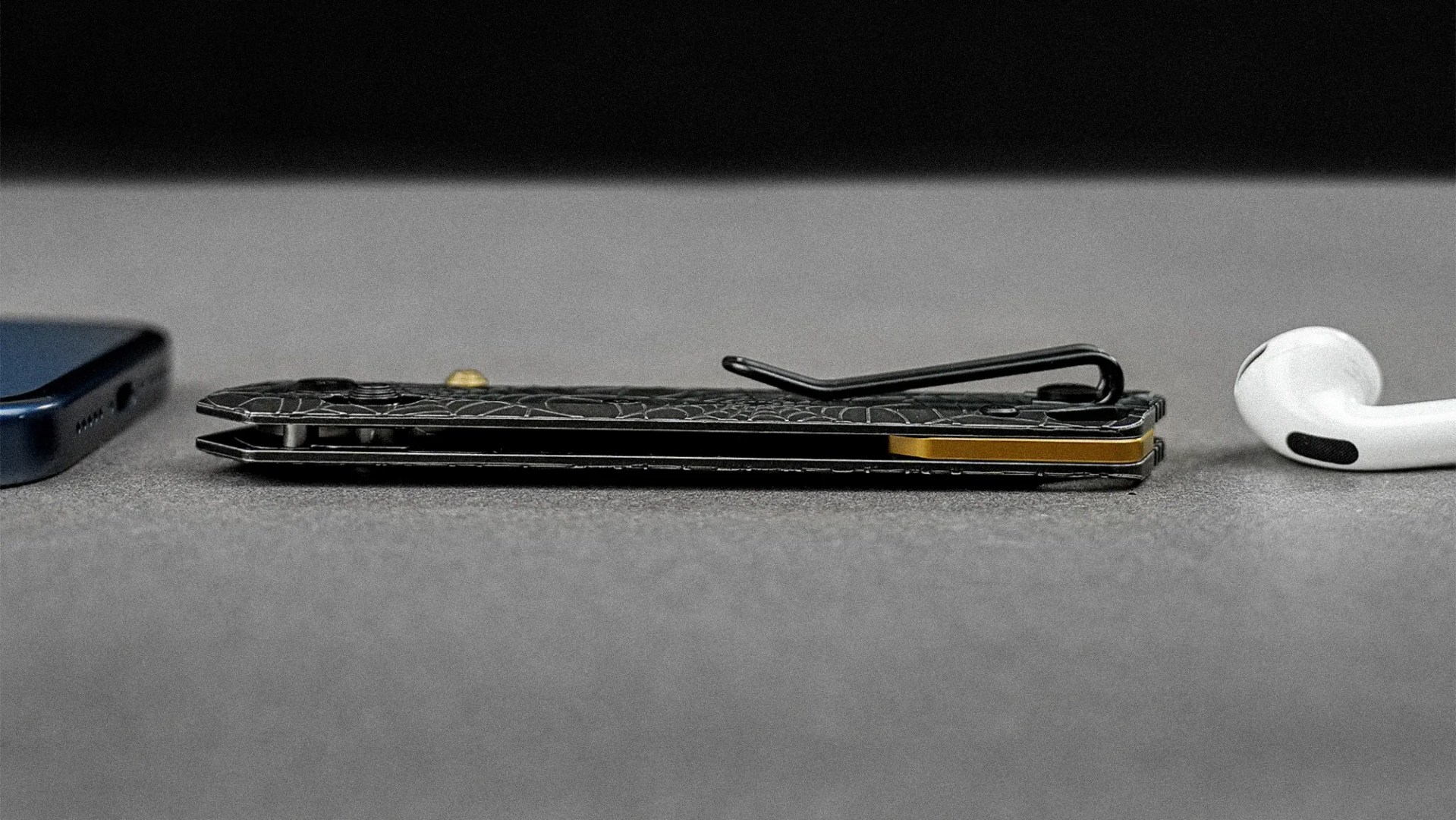 Black folding knife with a textured handle and gold accent lying flat between a smartphone and a white wireless earbud on a gray surface.