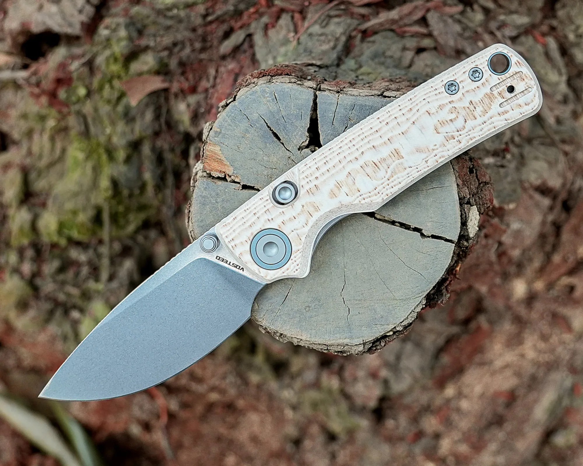 Folding knife with a stonewashed blade and white textured handle resting on a tree stump.