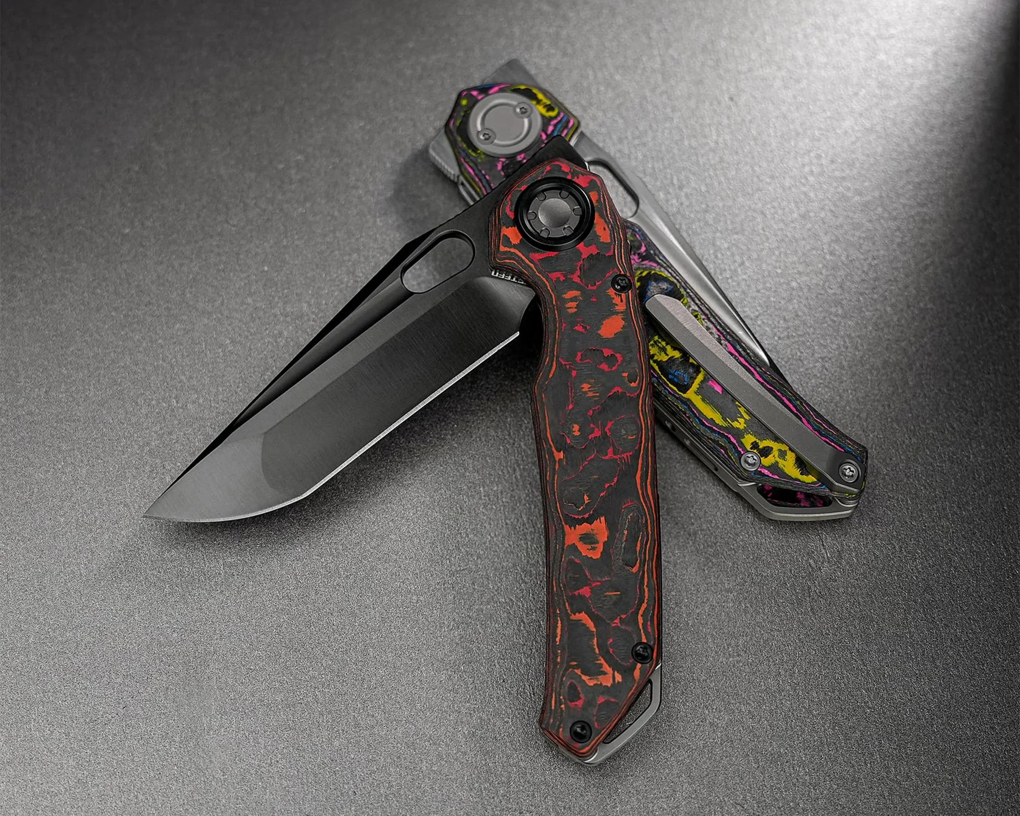 Two folding knives with black blades and colorful patterned handles, one with red and black and the other with yellow, pink, and blue, on a gray surface.