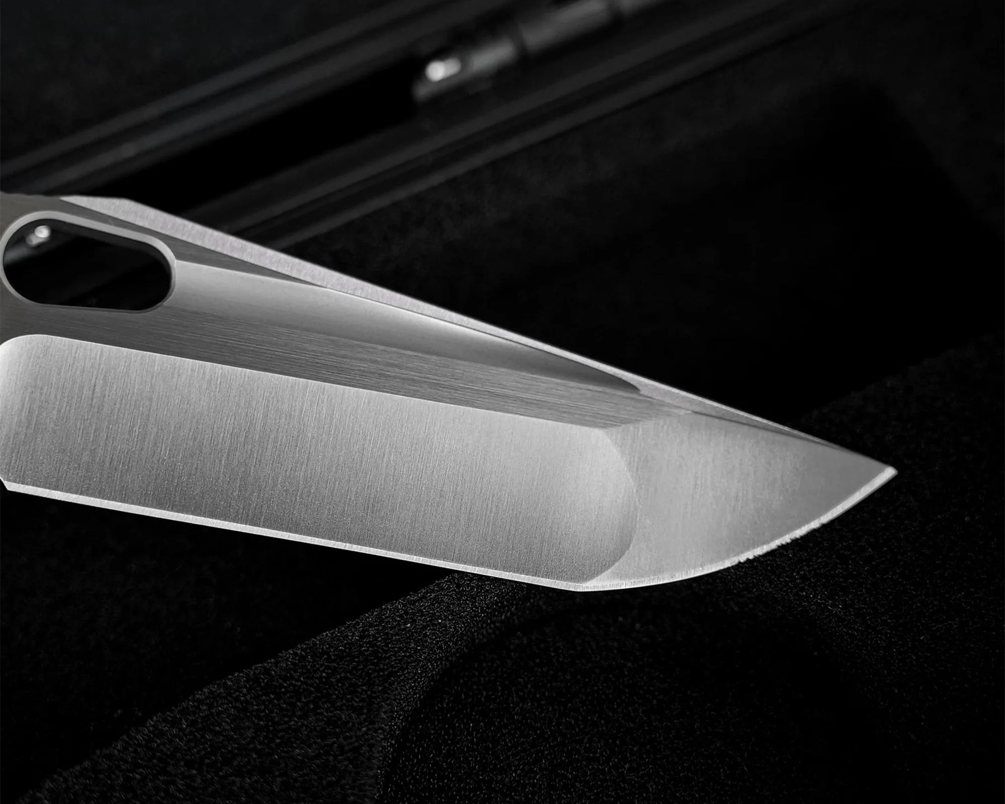 Close-up of a sharp, silver knife blade with a hole near the handle, resting on a black textured surface.