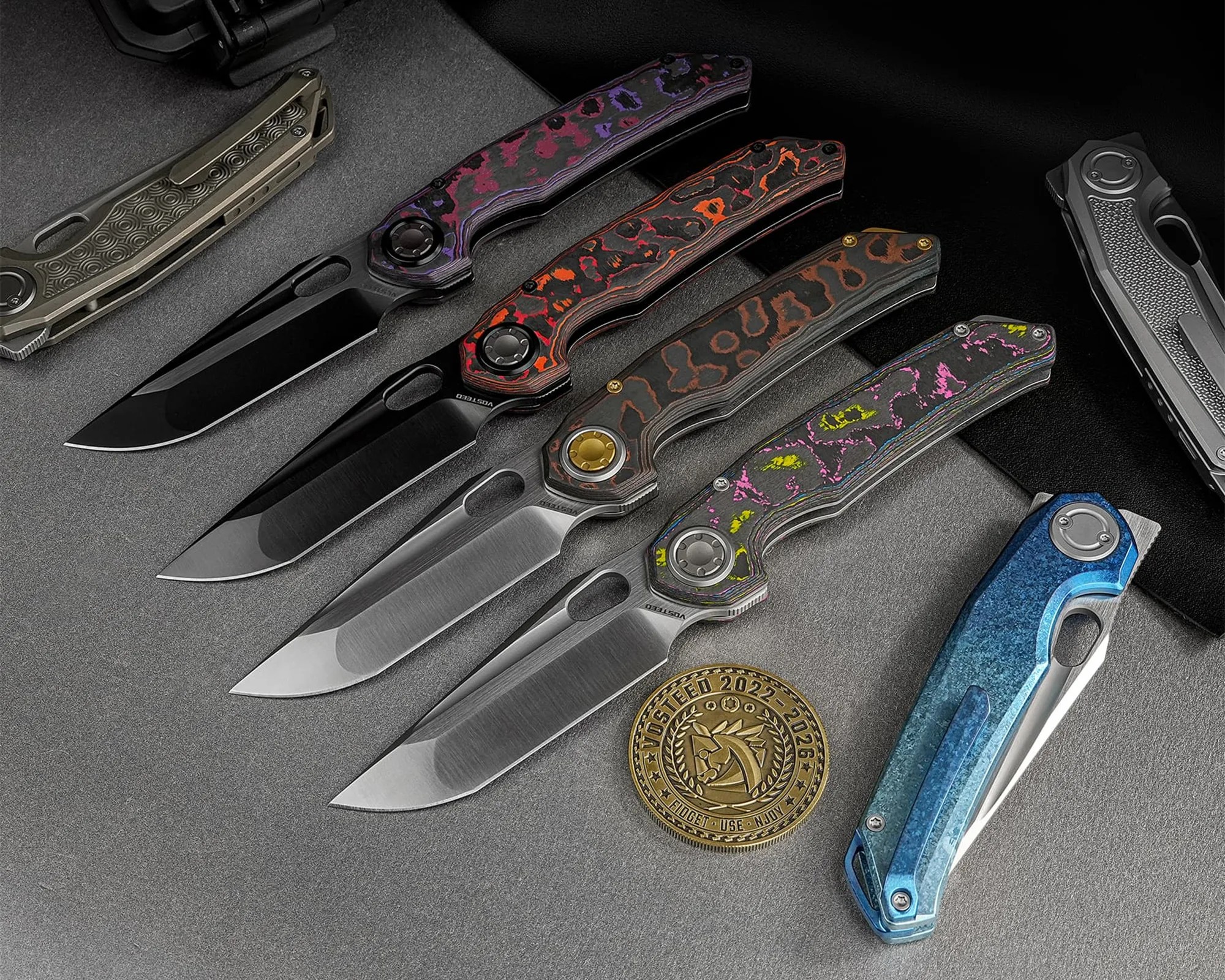 Five folding knives with patterned handles in purple, orange, brown, pink, and blue, arranged on a gray surface next to a gold coin.