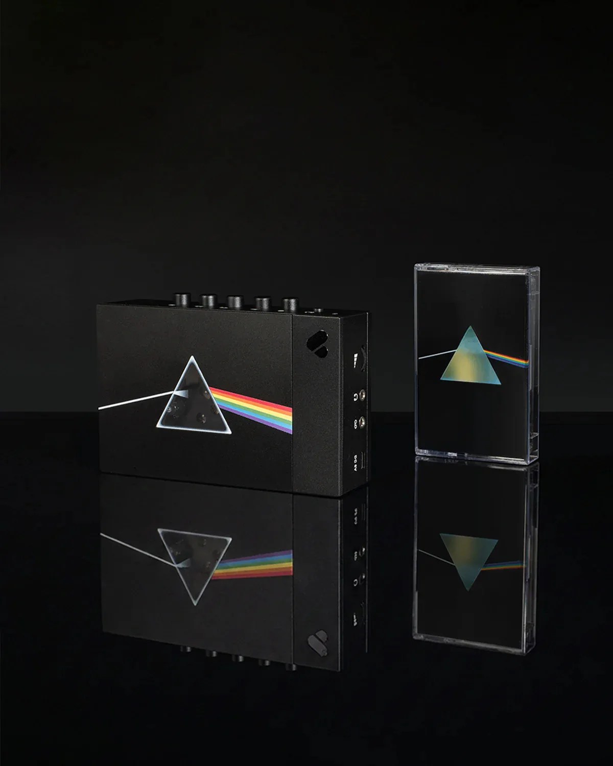 Black rectangular audio device and cassette tape case with prism and rainbow design on a reflective black surface.