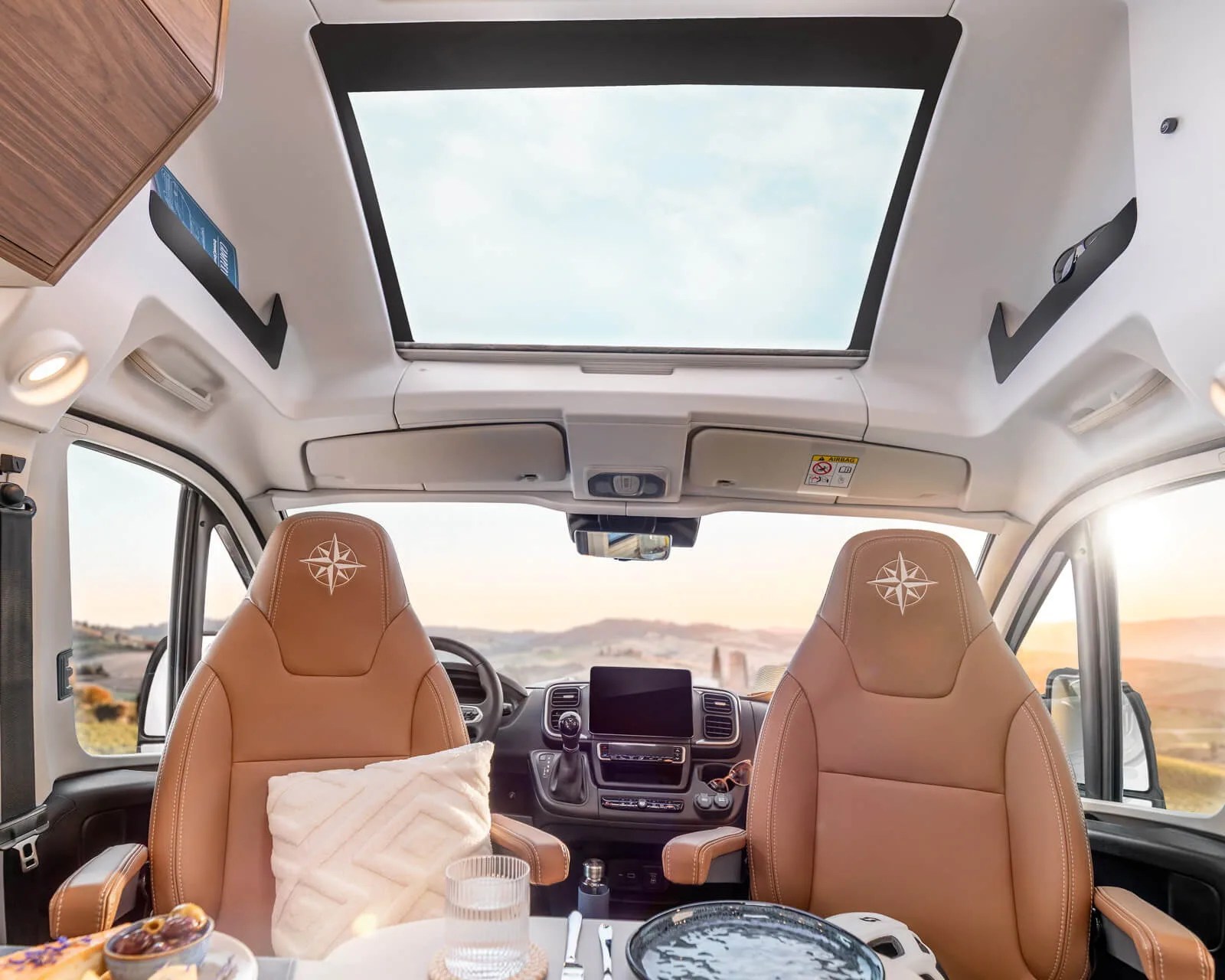 Interior of a vehicle with two tan leather seats featuring compass logos, a large sunroof, and a table set with dishes and a glass.