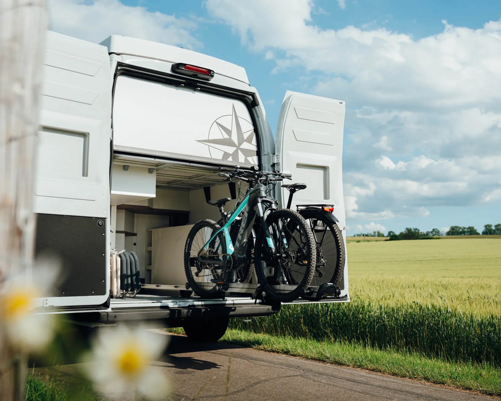 Two electric mountain bikes mounted on the back of a white camper van with open rear doors, parked by a grassy field.