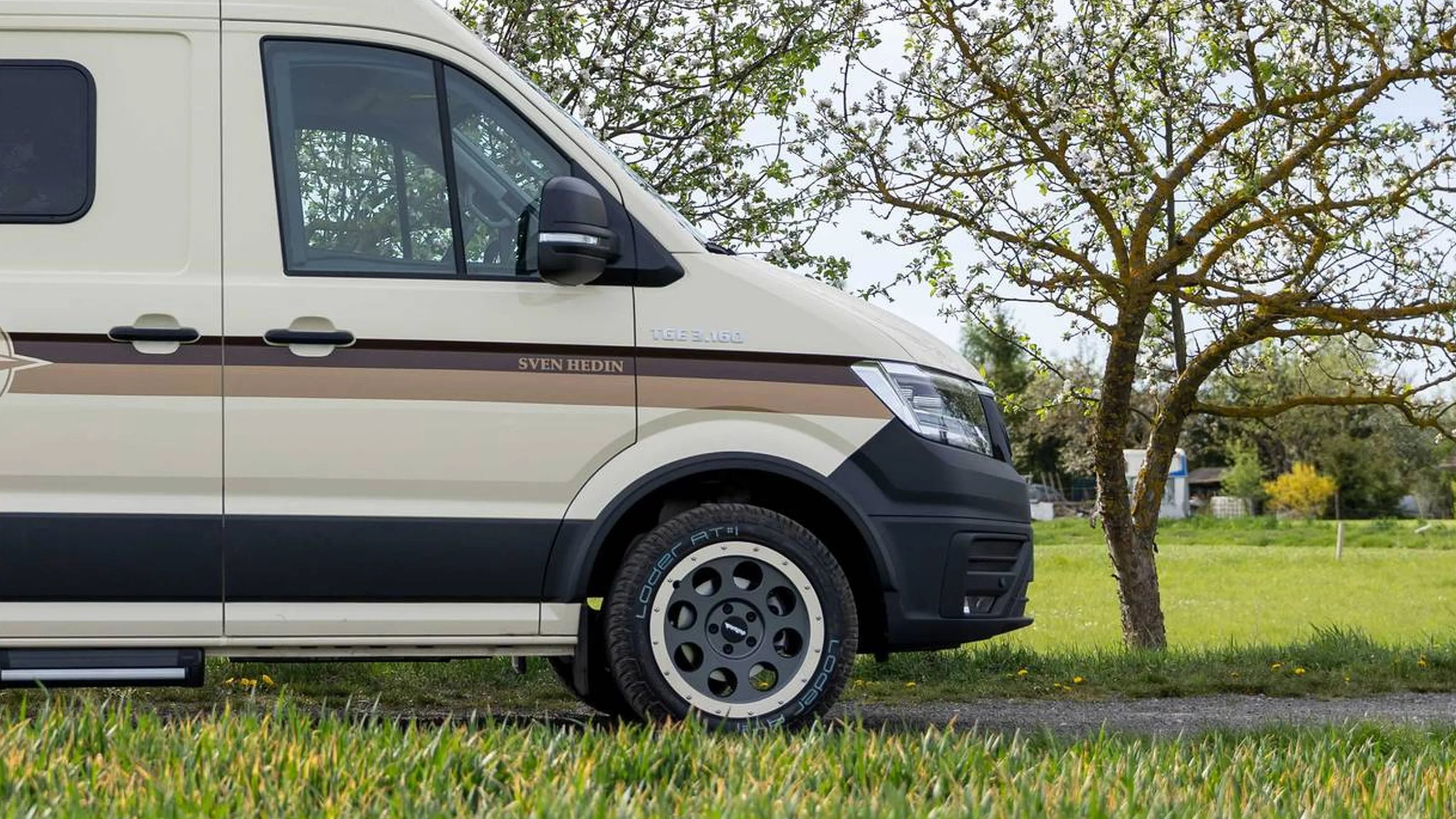 Beige camper van with brown stripes and black trim parked on grass near a tree.