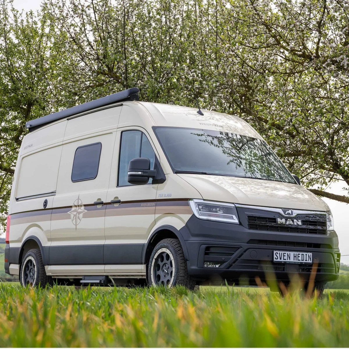 Beige MAN camper van with black trim and roof awning parked on grass under blooming trees.