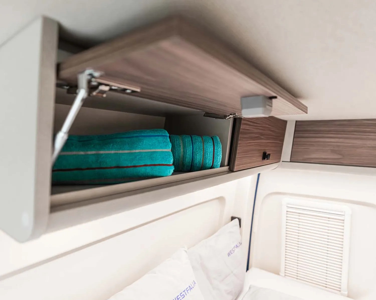 Overhead wooden cabinet with an open door revealing folded turquoise towels inside, above a bed with white pillows.