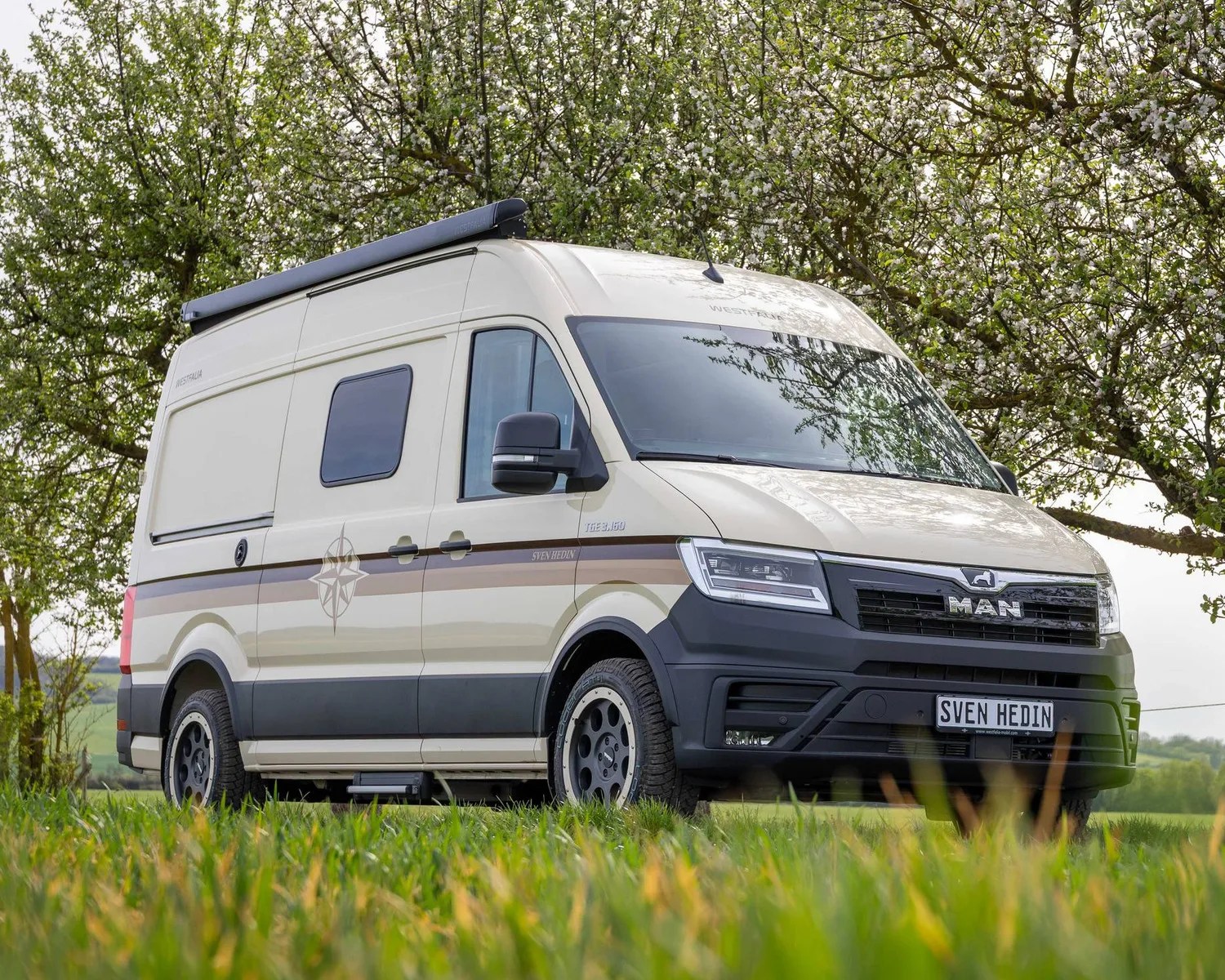 Beige MAN camper van with black trim and roof awning parked on grass under a tree with green leaves.