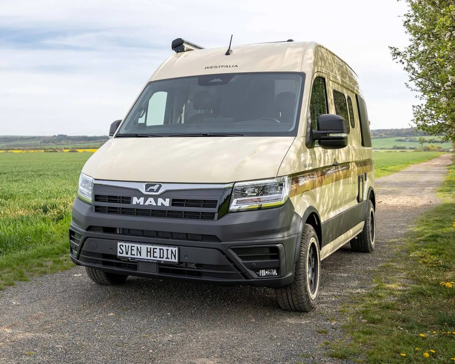 Beige MAN Westfalia camper van parked on a gravel path beside green fields and trees.