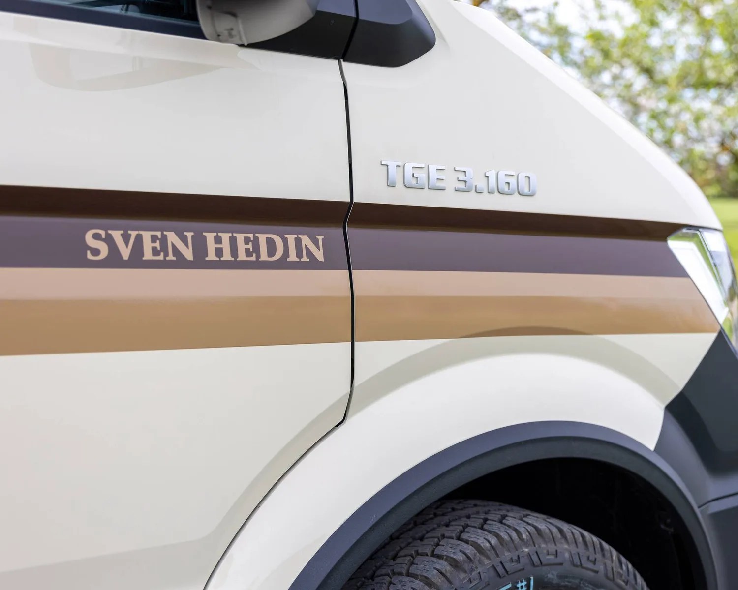 Close-up of a beige vehicle's side with brown stripes, "SVEN HEDIN" text, and "TGE 3.160" badge above the front wheel.