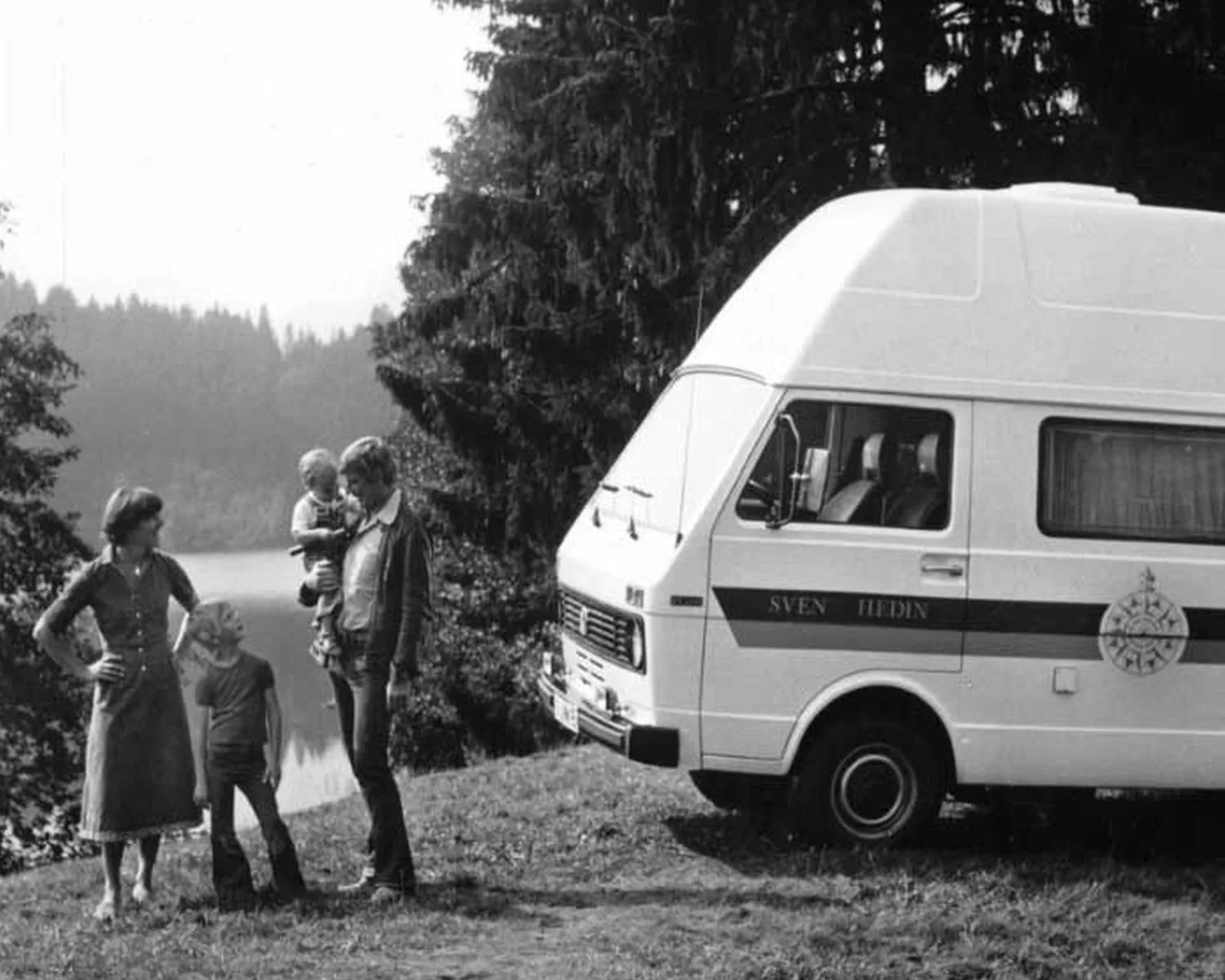 Black-and-white photo of a family of four standing on grass near a white camper van with "SVEN HEDIN" on the side by a lake and trees.