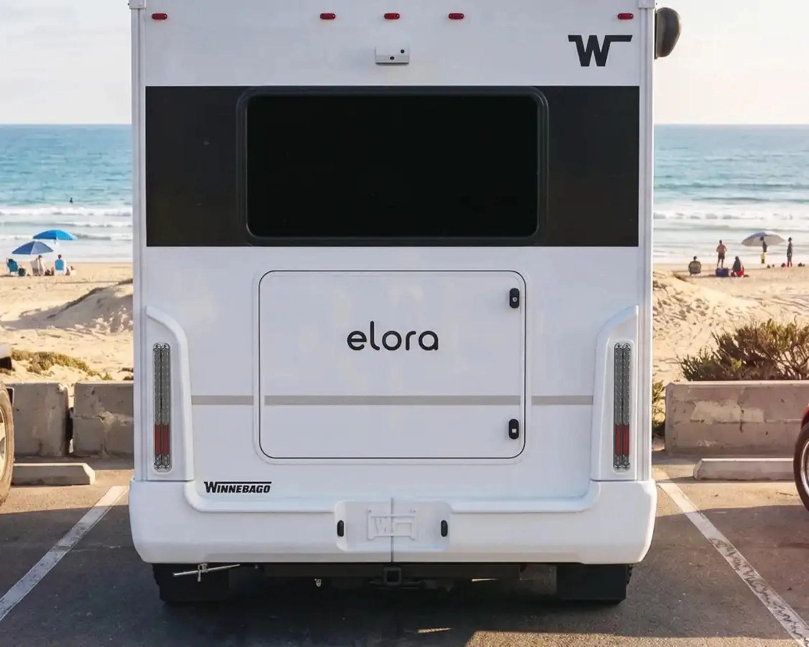 Rear view of a white Winnebago Elora RV parked near a beach with ocean and people in the background.
