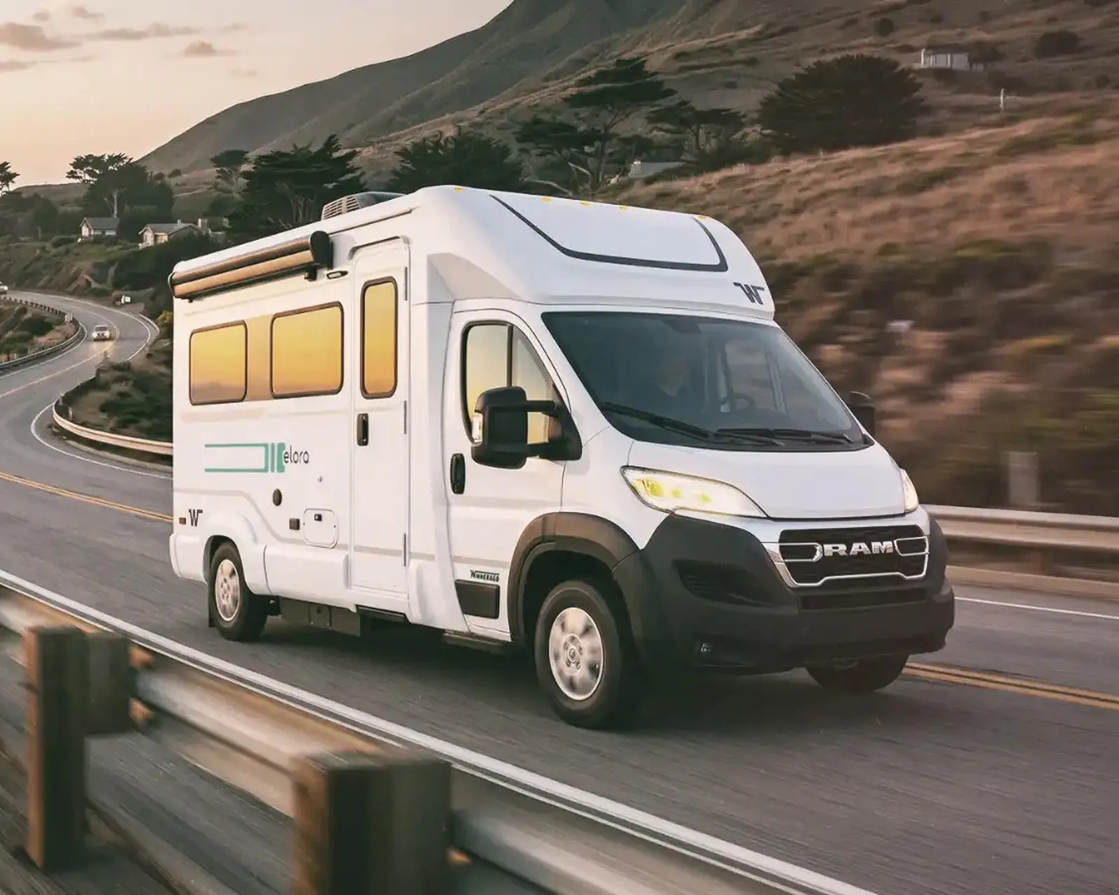 White RAM motorhome with tinted windows driving on a winding road near hills at sunset.