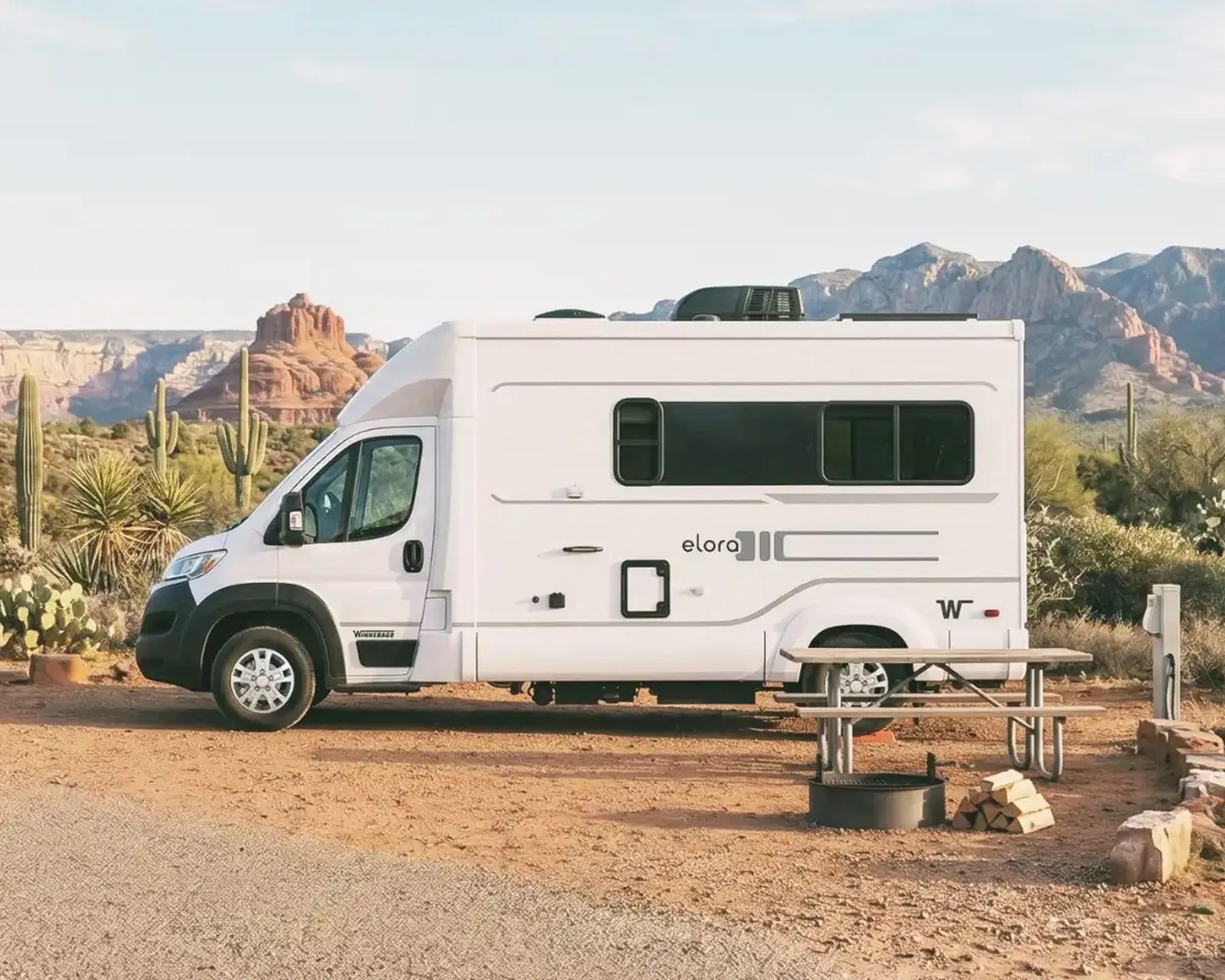 White Winnebago Elora camper van parked in a desert campsite with a picnic table and fire pit nearby.