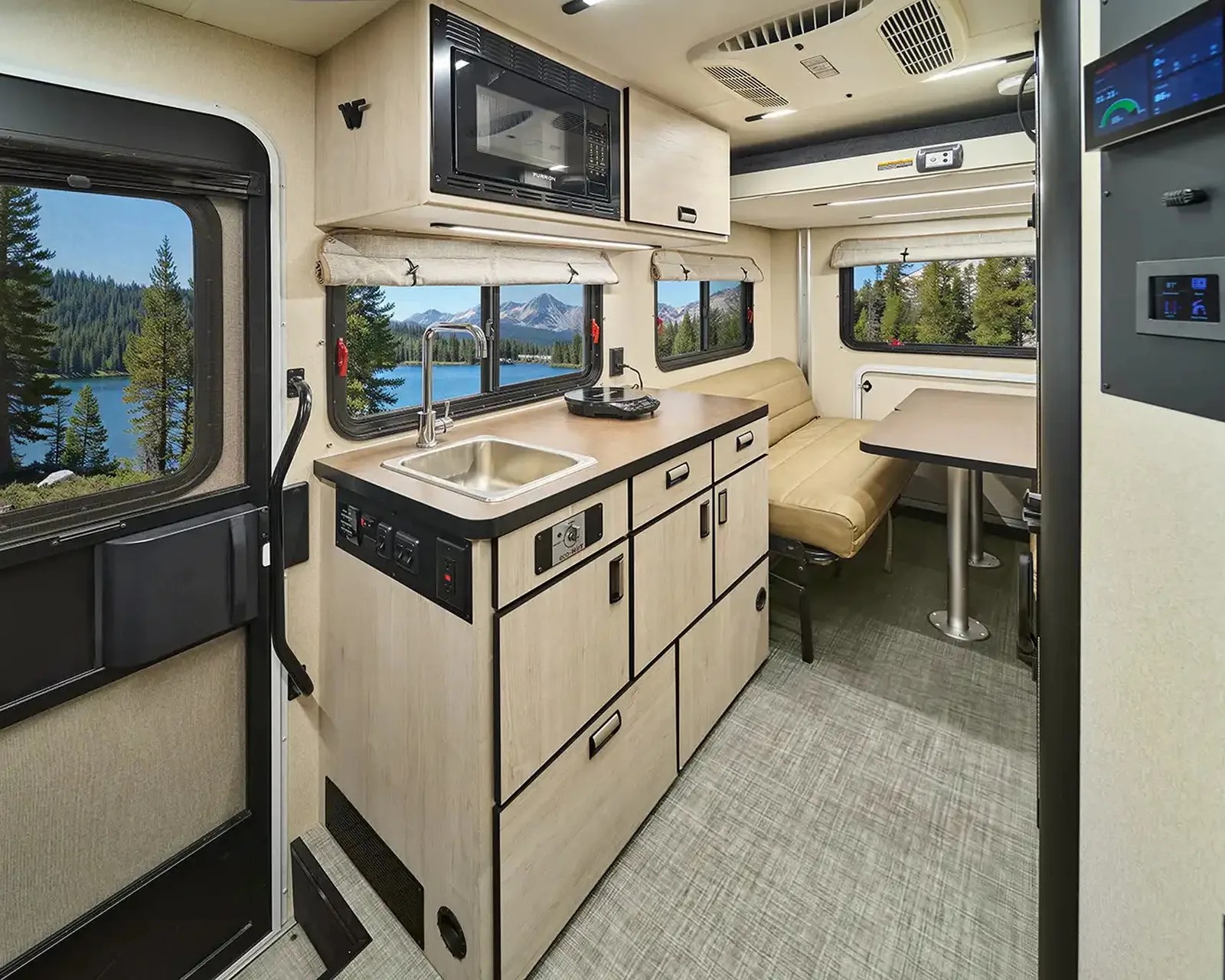 Compact RV interior kitchen with light wood cabinets, stainless steel sink, microwave, beige cushioned bench, and table by windows showing forest and lake.